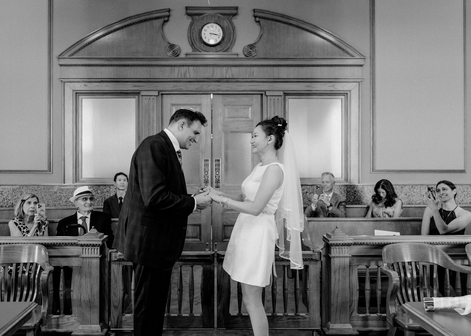 Black and white photo of a couple exchanging rings at a courthouse wedding ceremony with guests seated behind them.
