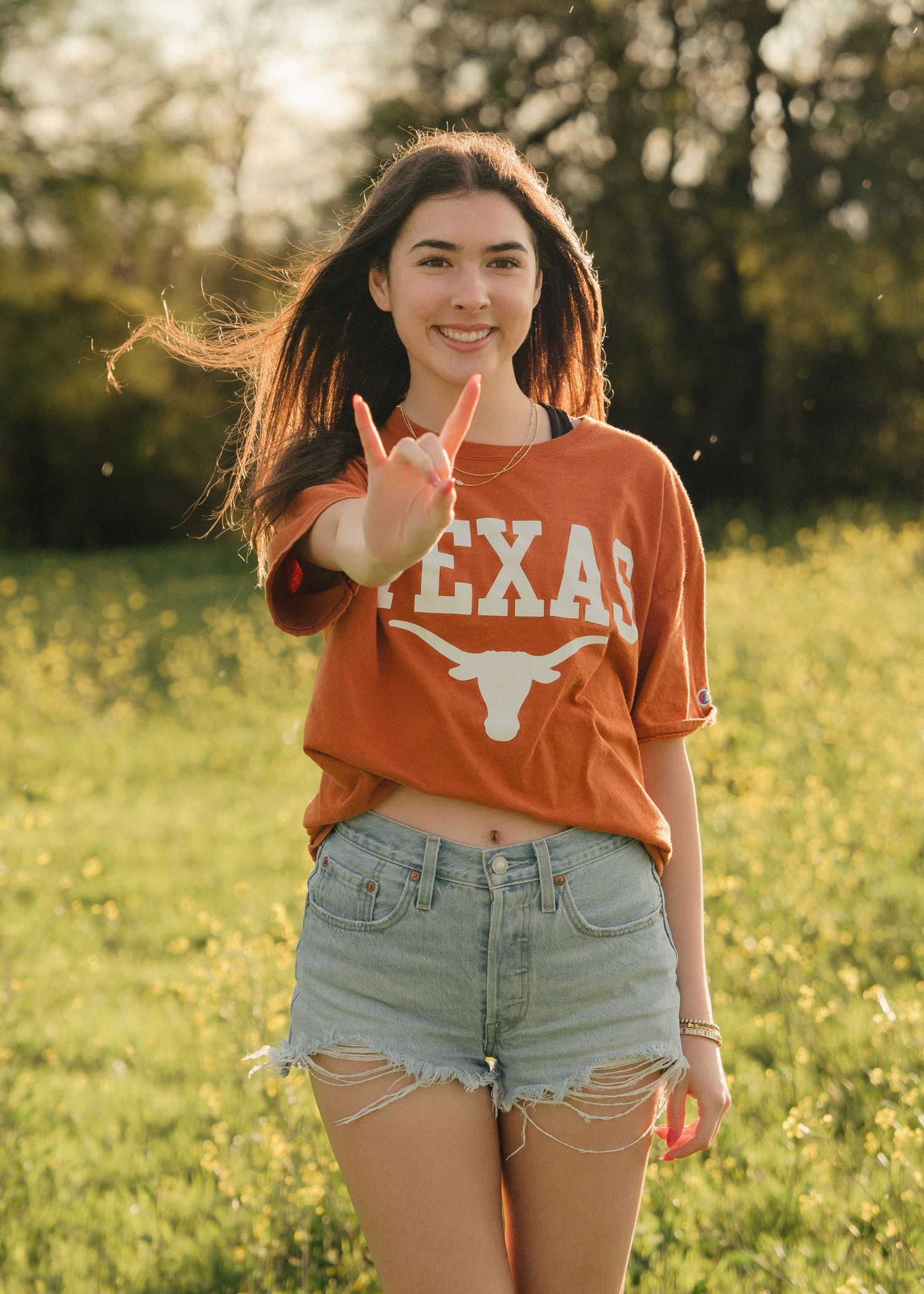 Young woman in burnt orange Texas Longhorns tee and denim shorts making hook em horns sign in sunny field.
