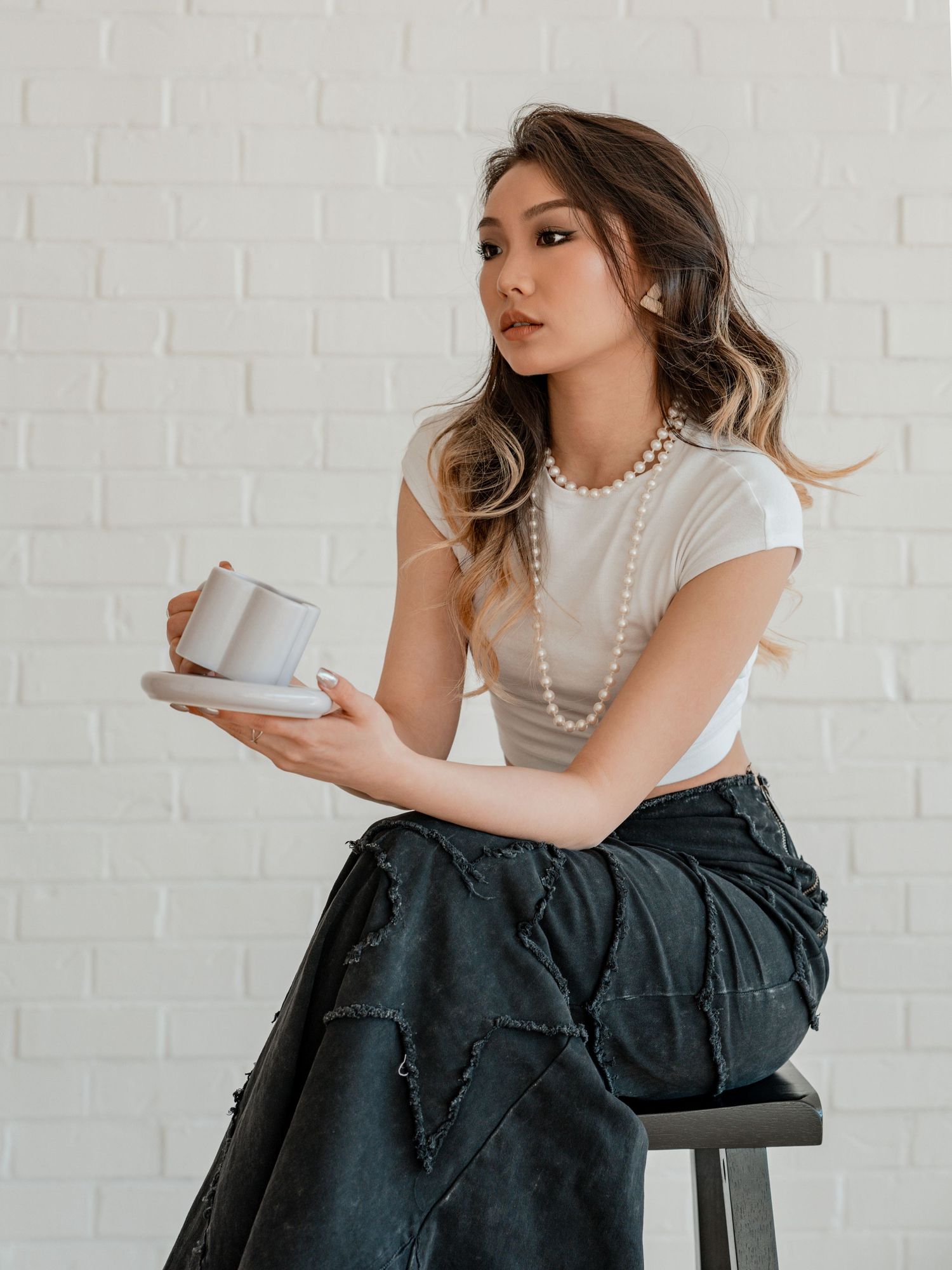 Young woman in white crop top, pearl necklace, and dark skirt sits on stool holding a cup and saucer.
