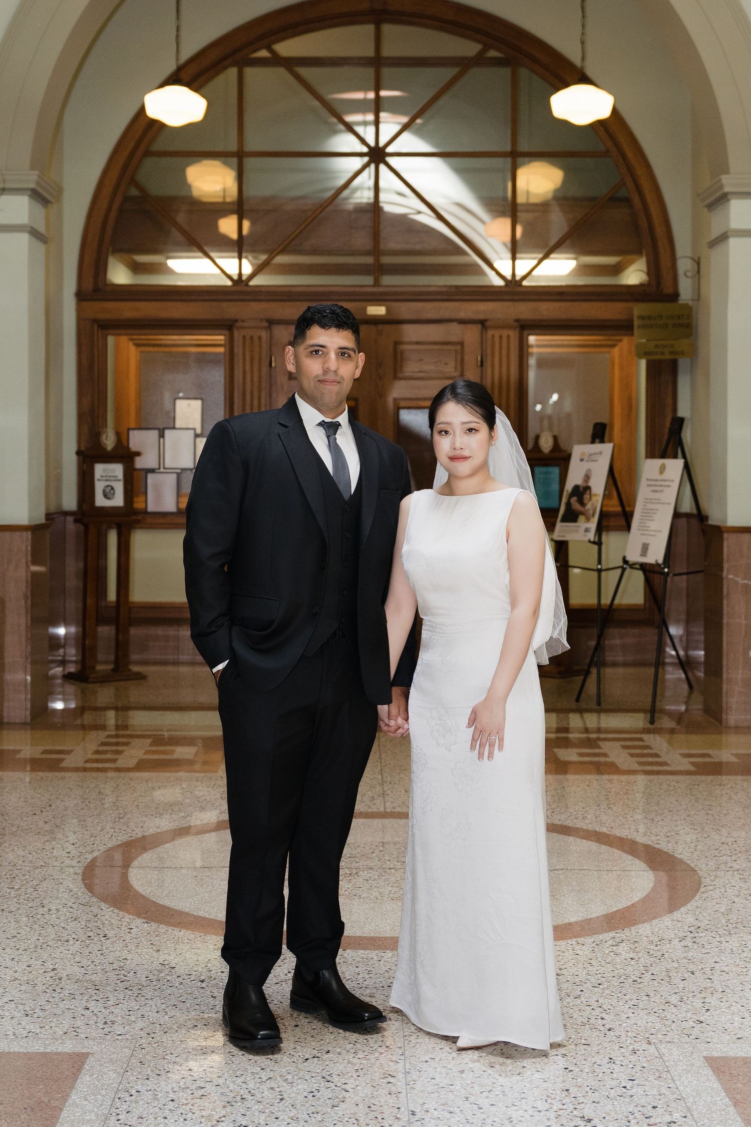 Bride and groom posing in elegant building lobby, she in white gown with veil, he in black suit.