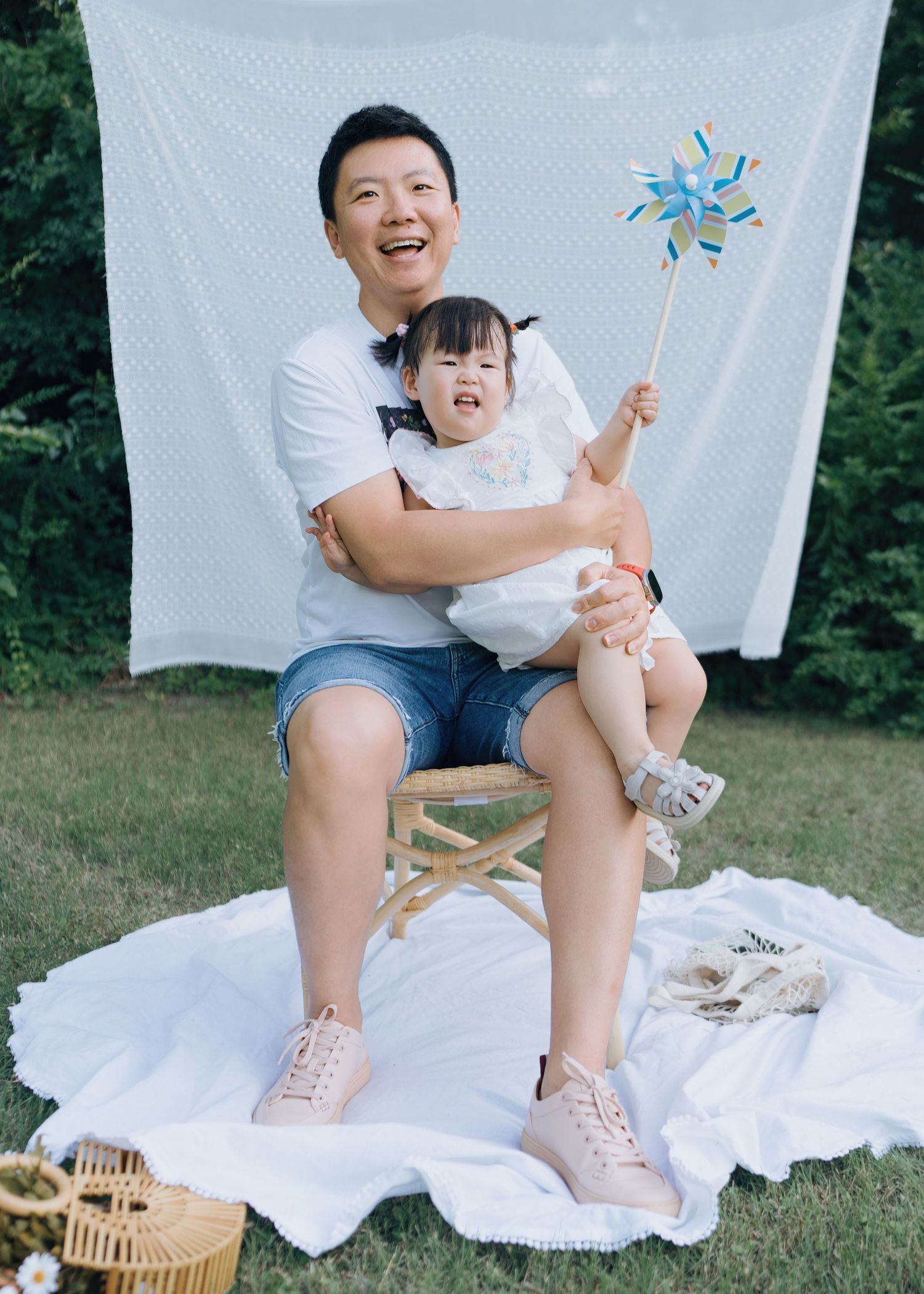 A laughing Asian grandfather sits on a chair outdoors holding a toddler with a colorful pinwheel.