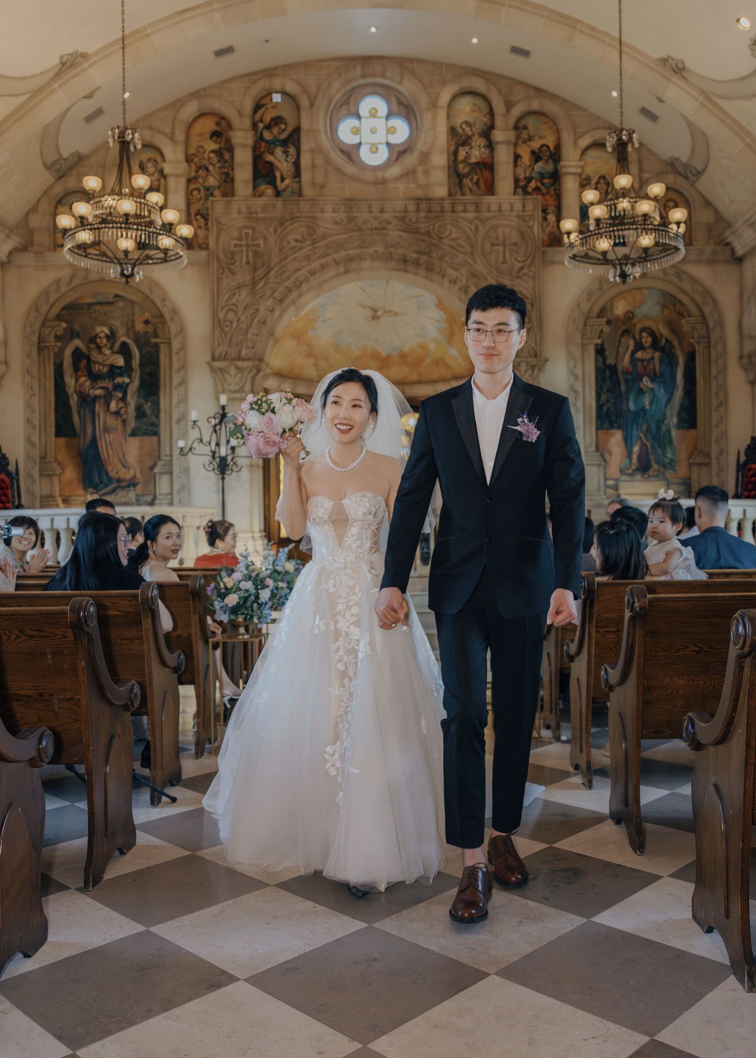 Smiling bride and groom walk hand in hand down the aisle of a beautifully decorated chapel.
