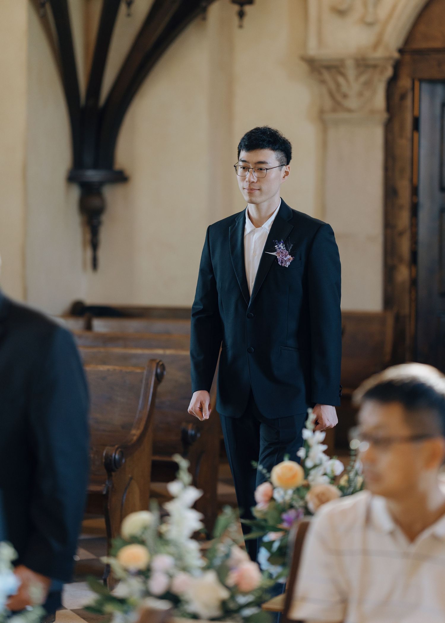Dapper groom in black suit with floral boutonniere walks down church aisle during wedding ceremony.