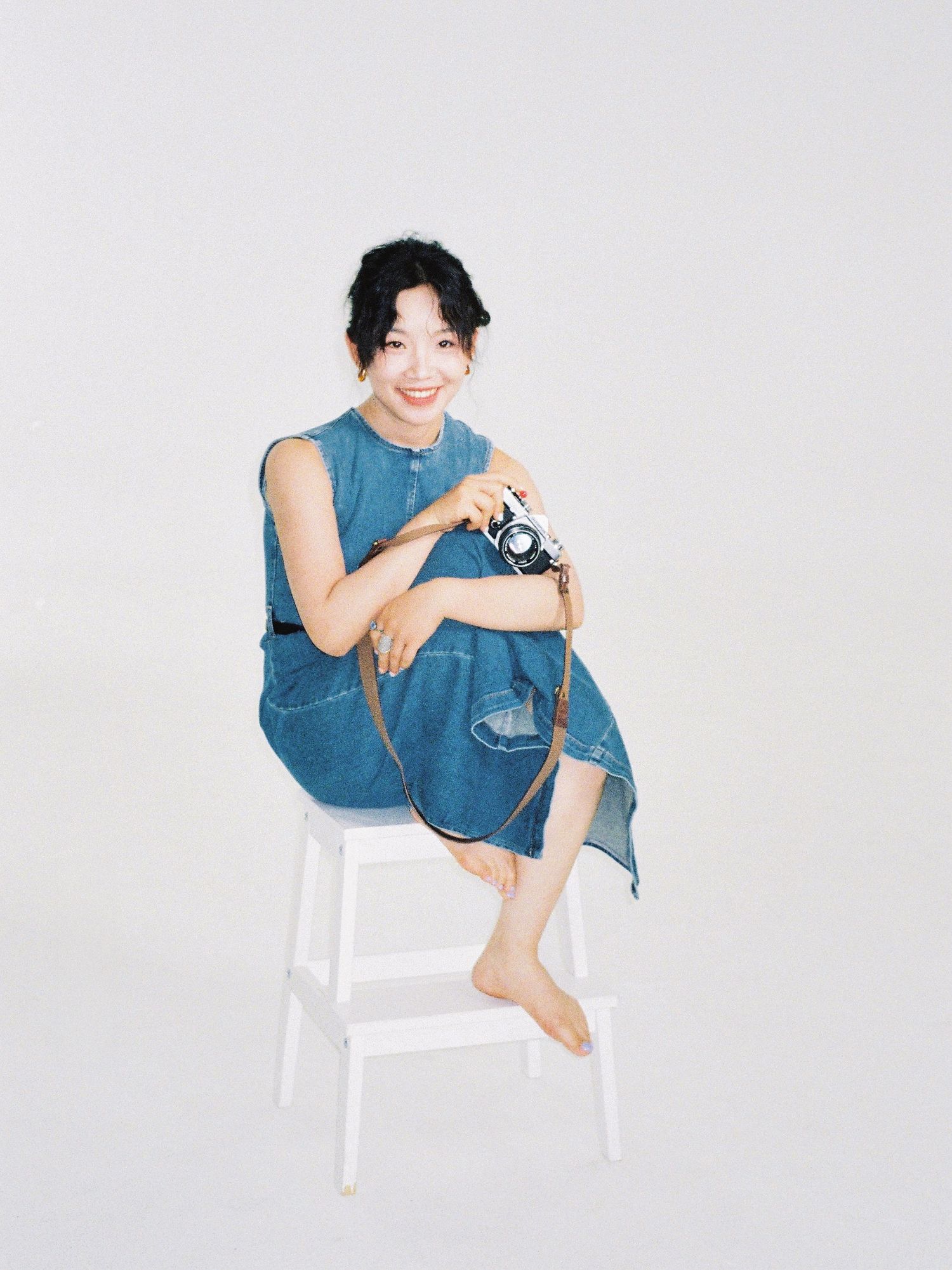 Smiling young woman in denim dress sitting on white stool holding a camera, barefoot against light background.