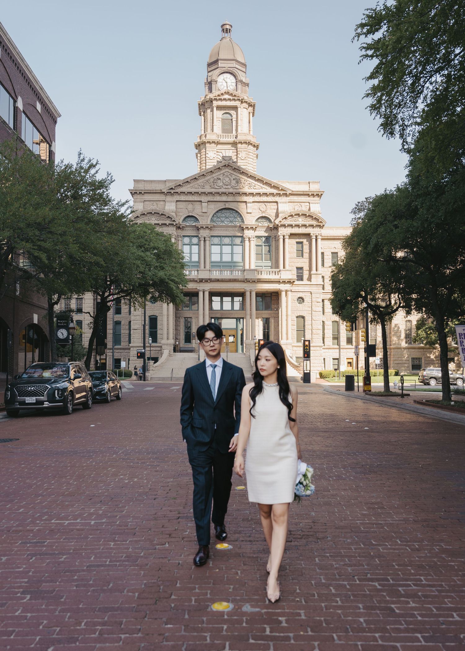 Couple in wedding attire walking in front of a historic courthouse on a brick plaza.