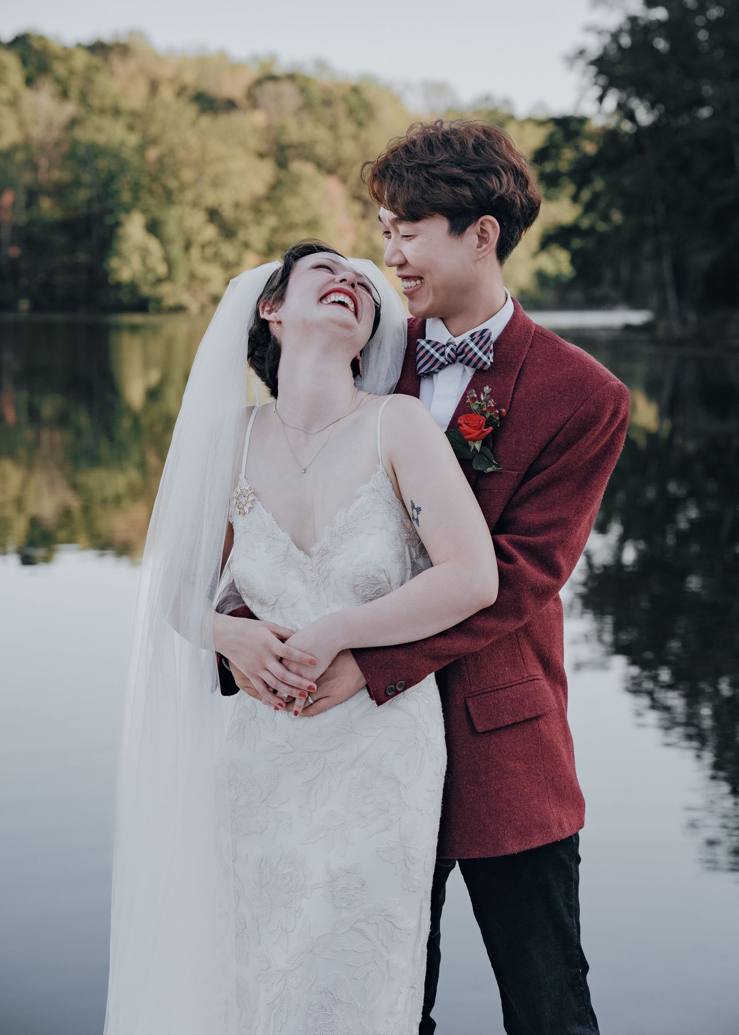 Groom in red suit embraces laughing bride in white lace gown by a peaceful lake.