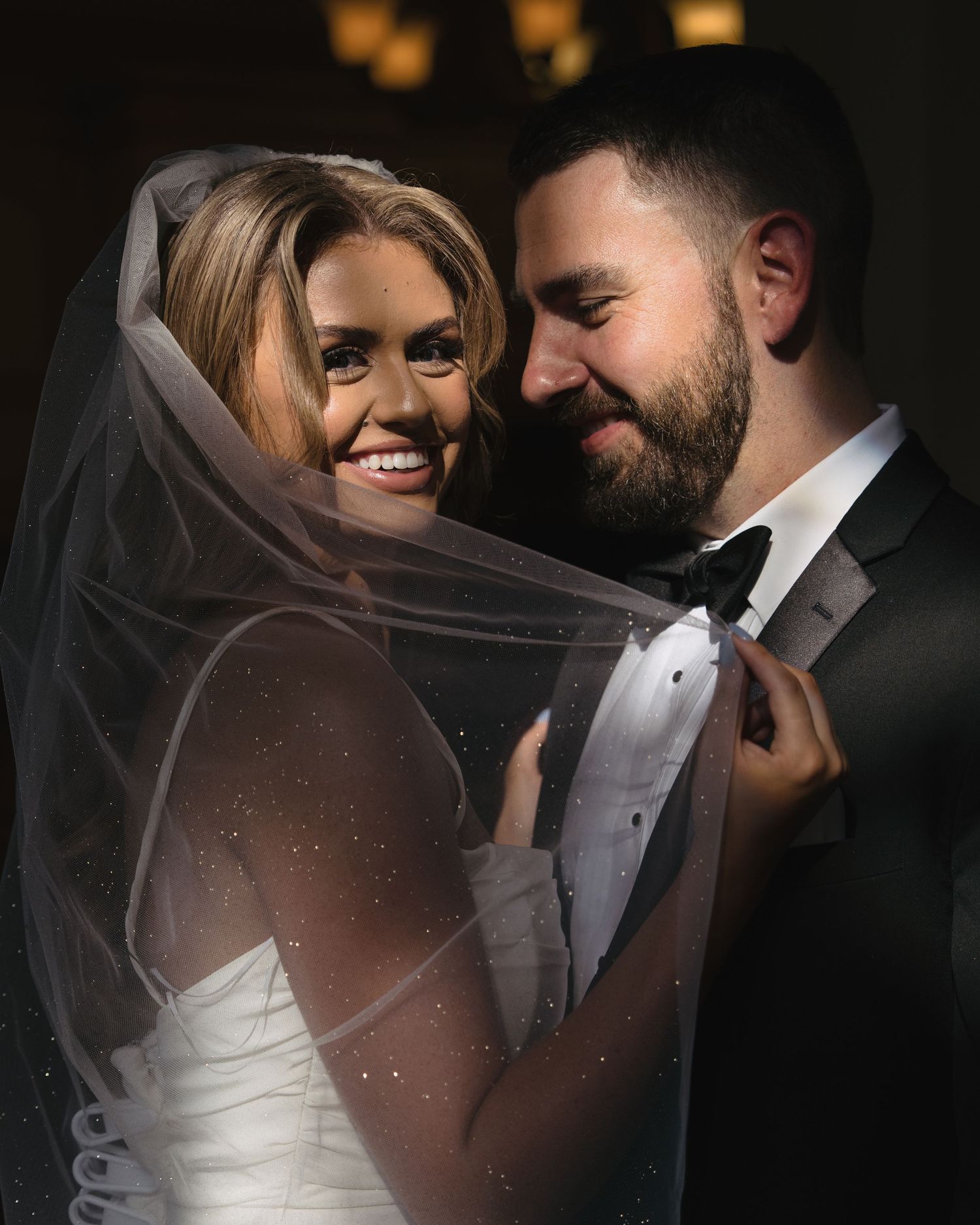 Smiling bride in white gown with veil embracing bearded groom in black tuxedo indoors.