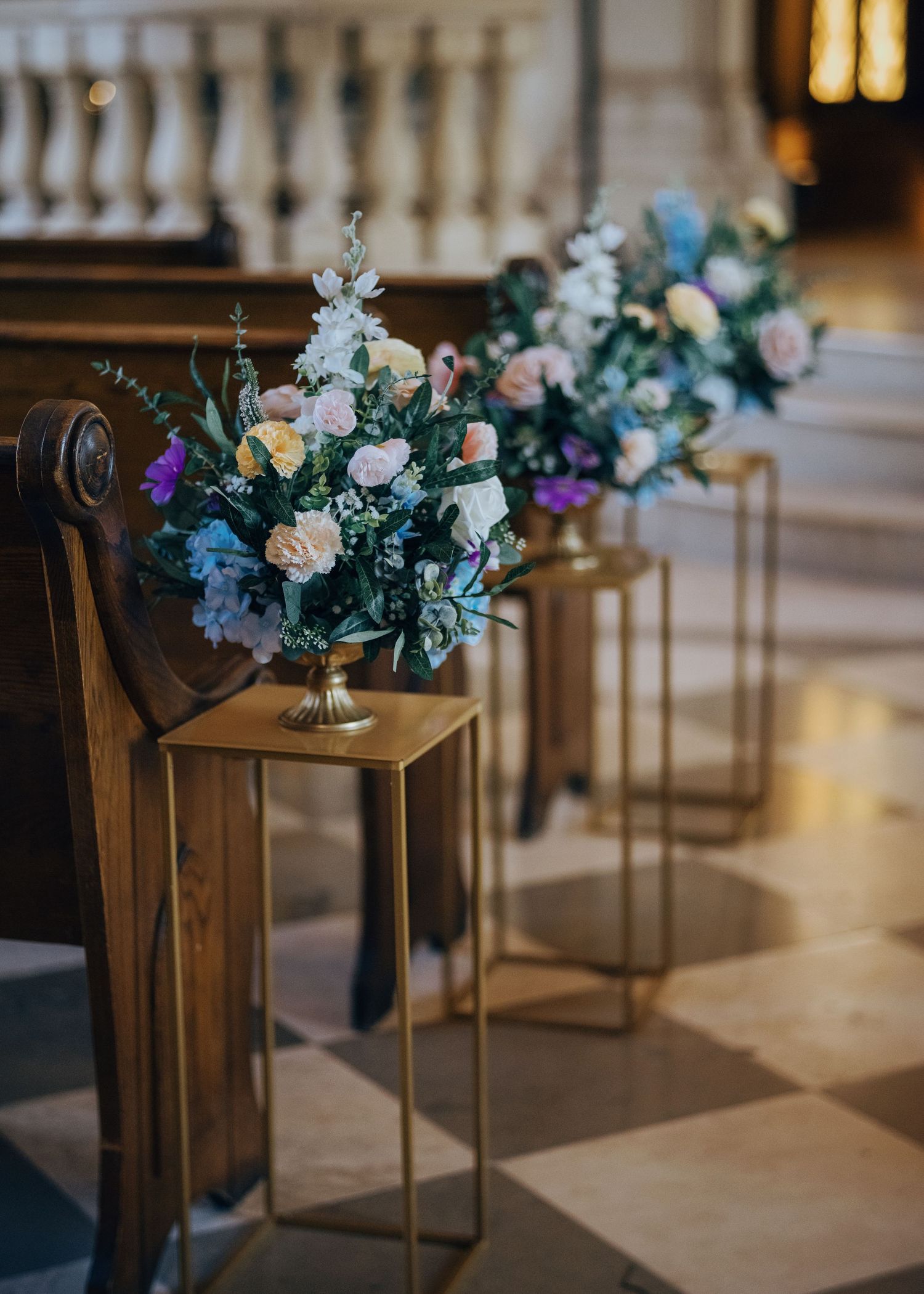 Elegant wedding aisle decor with pastel floral arrangements in gold vases beside wooden church pews.