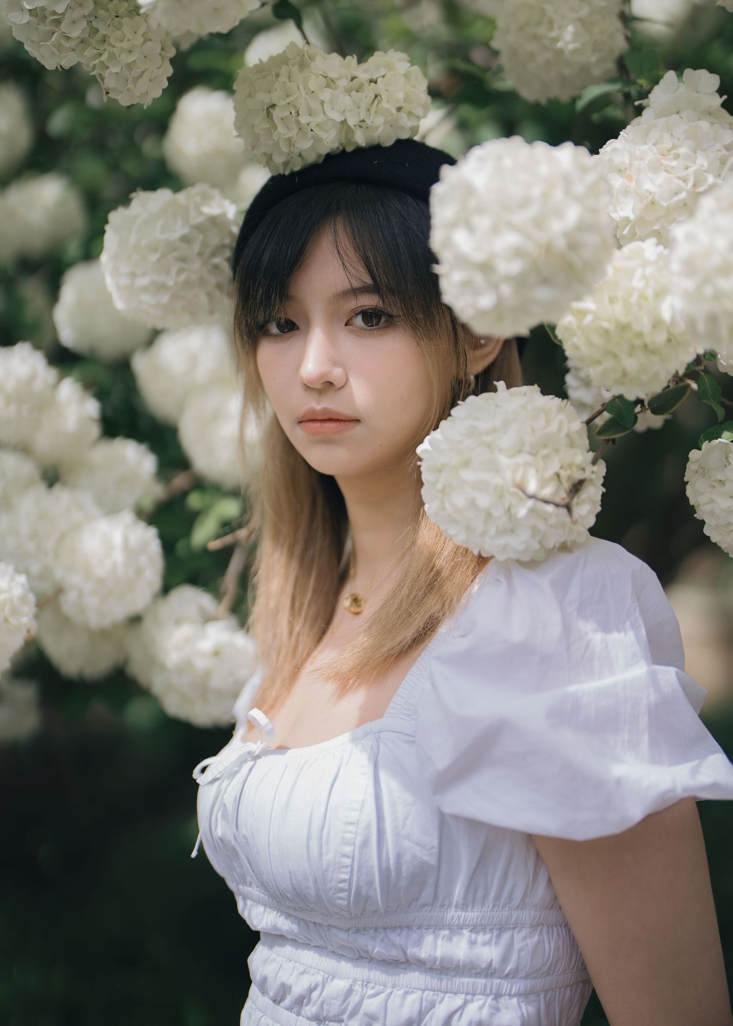 Young Asian woman in white puff sleeve dress surrounded by large white hydrangea blooms outdoors.