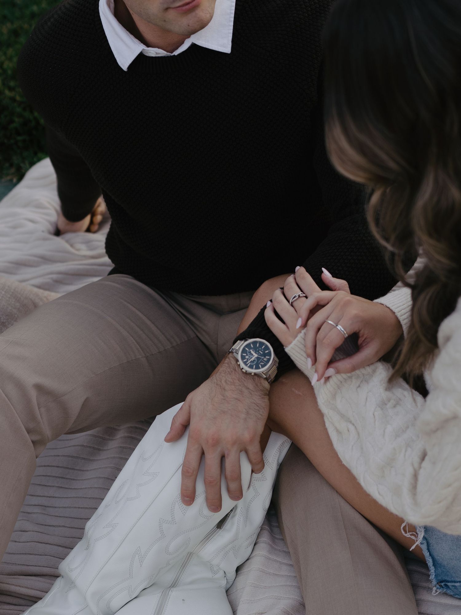 Close-up of couple holding hands on a blanket, showing engagement ring and stylish watch.