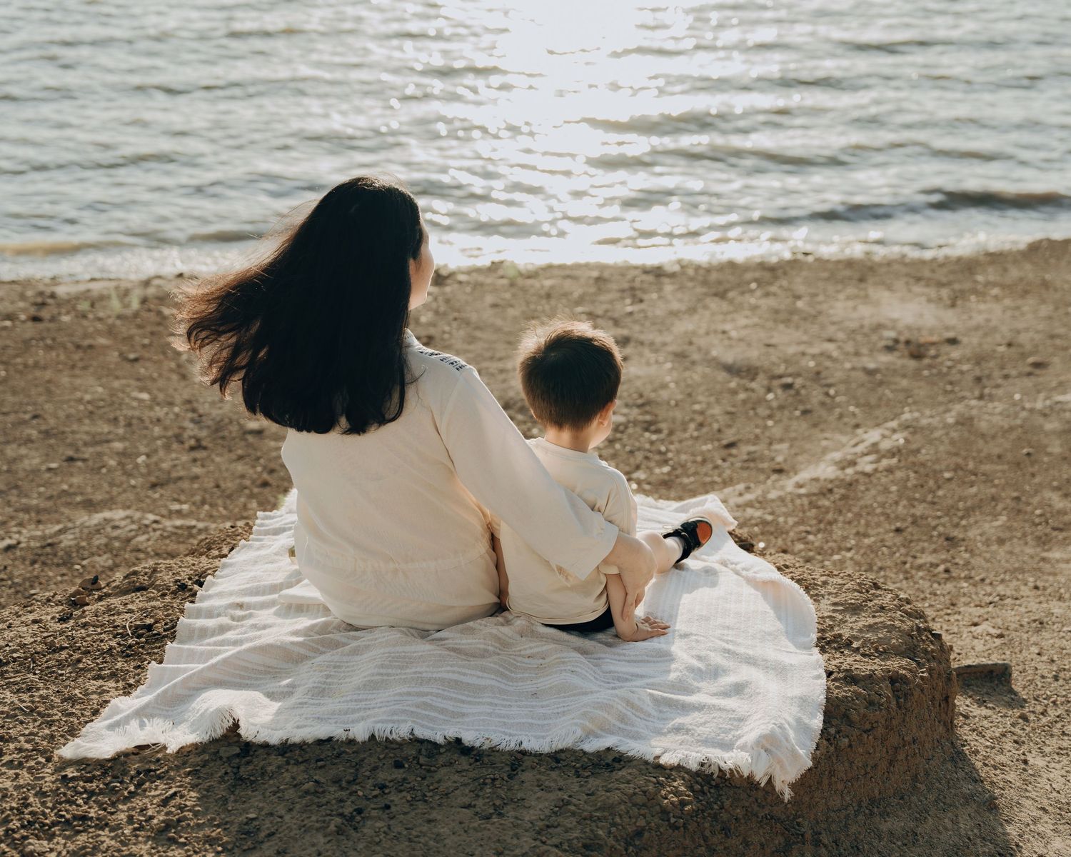 Mother and toddler sit together on a white blanket by the water enjoying a calm beachside moment.