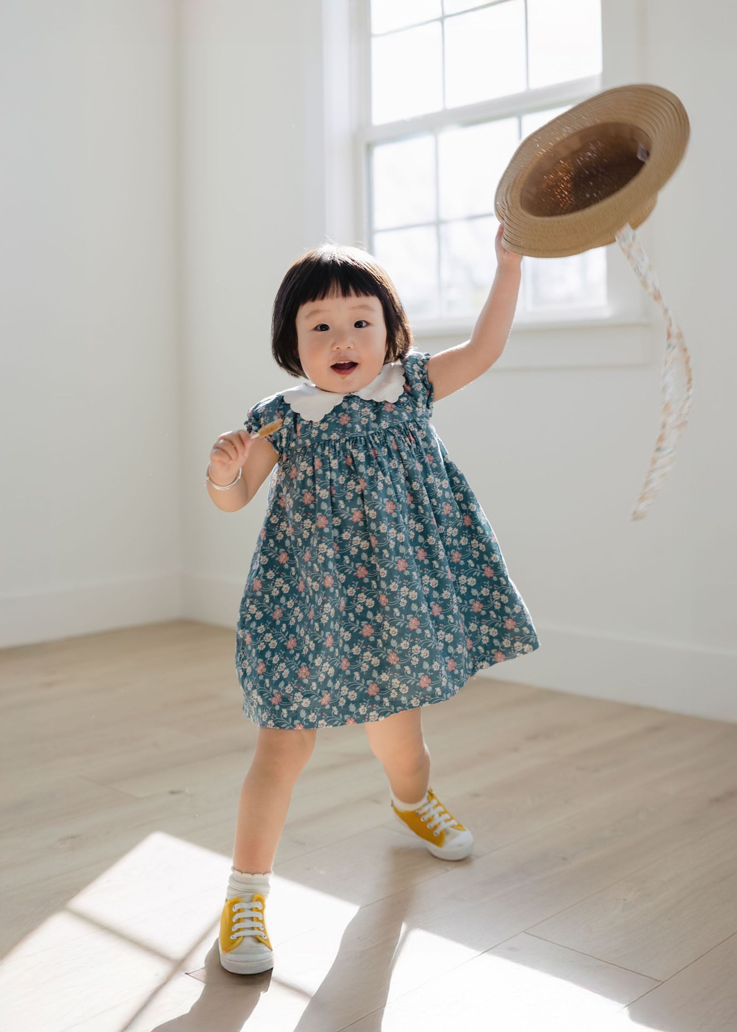 Happy toddler girl in teal floral dress joyfully waving straw hat in sunlit room.