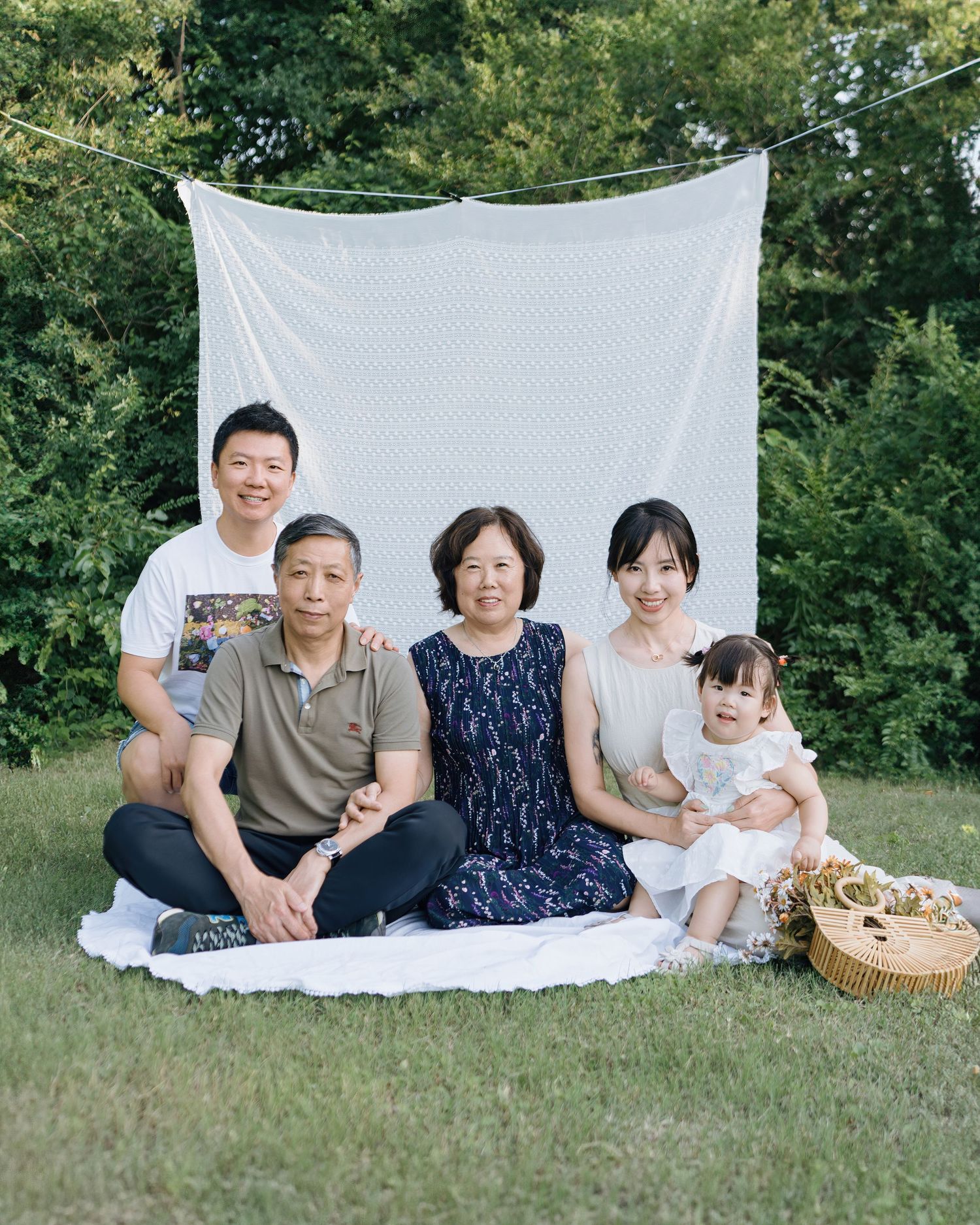 Asian multigenerational family of five posing together on lawn with white fabric backdrop outdoors.