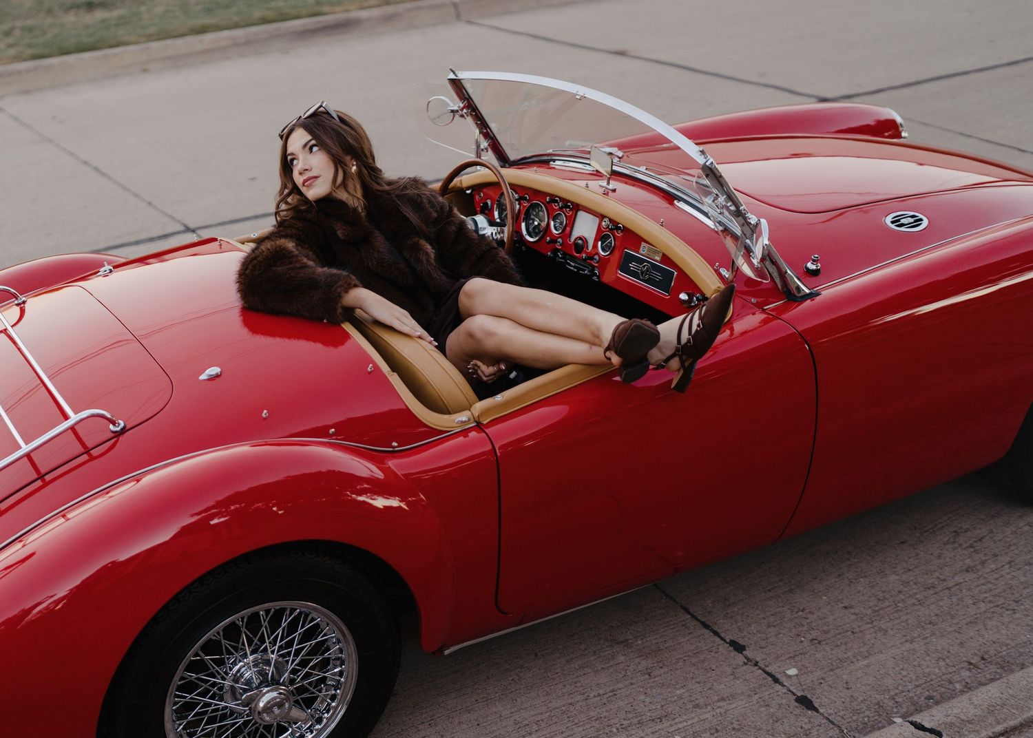 Woman in brown fur coat poses draped across red vintage MGA convertible sports car.