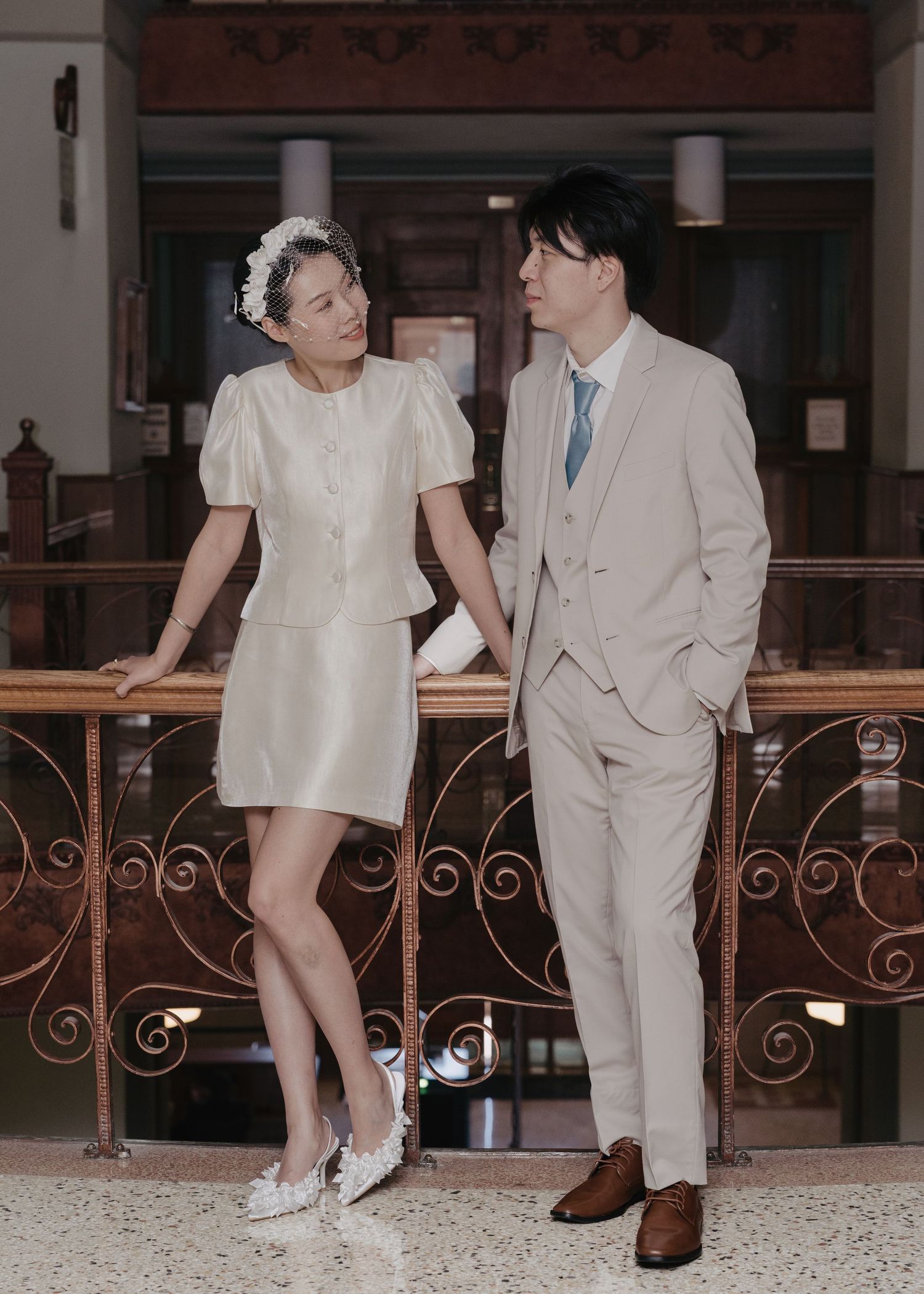 Couple in wedding attire posing by ornate railing, woman in white mini dress, man in beige suit with blue tie.