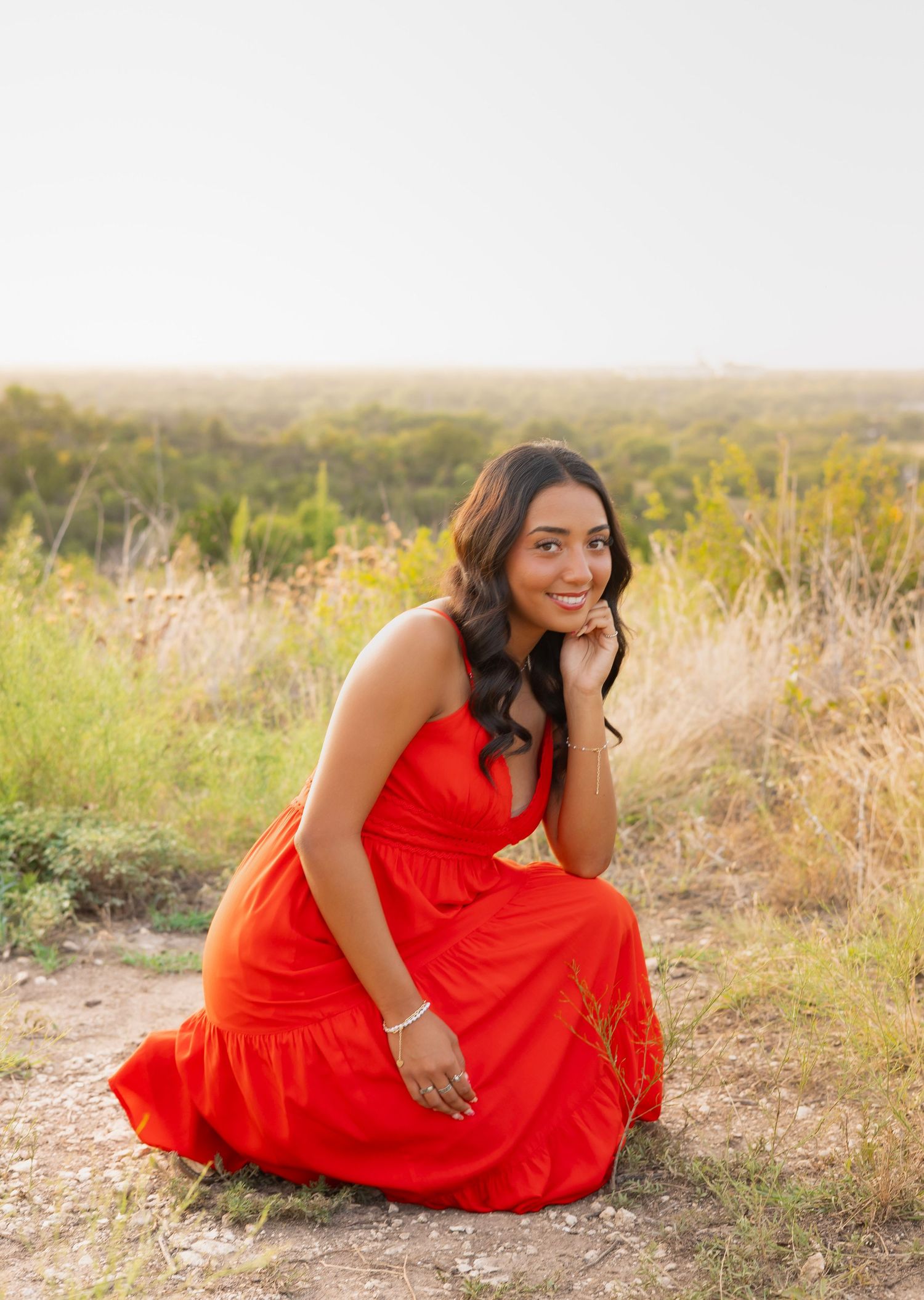 Cheerful woman in orange-red dress posing gracefully on a trail with open fields behind her.