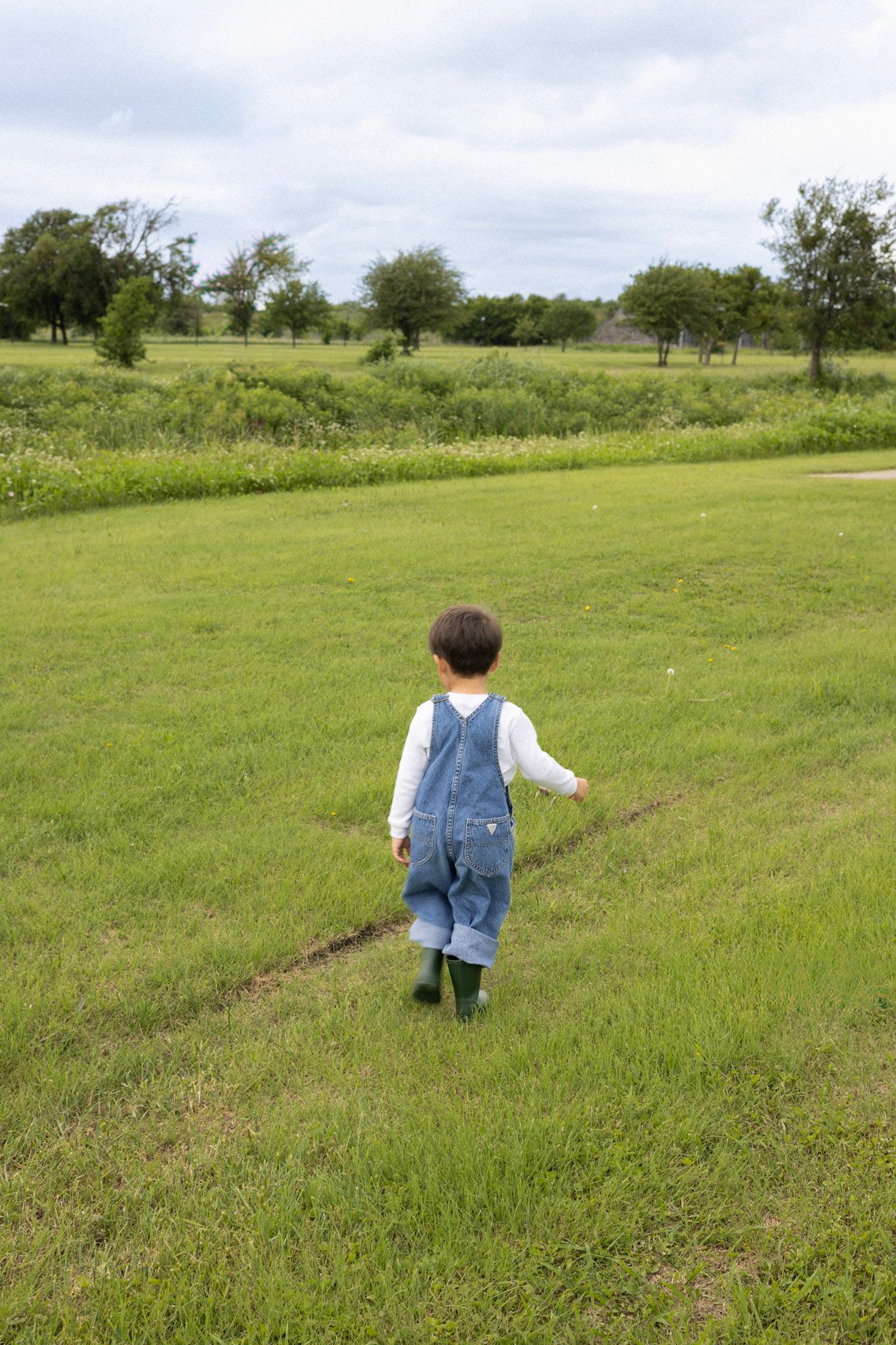 Young toddler in denim overalls and green boots walking across a lush green field.