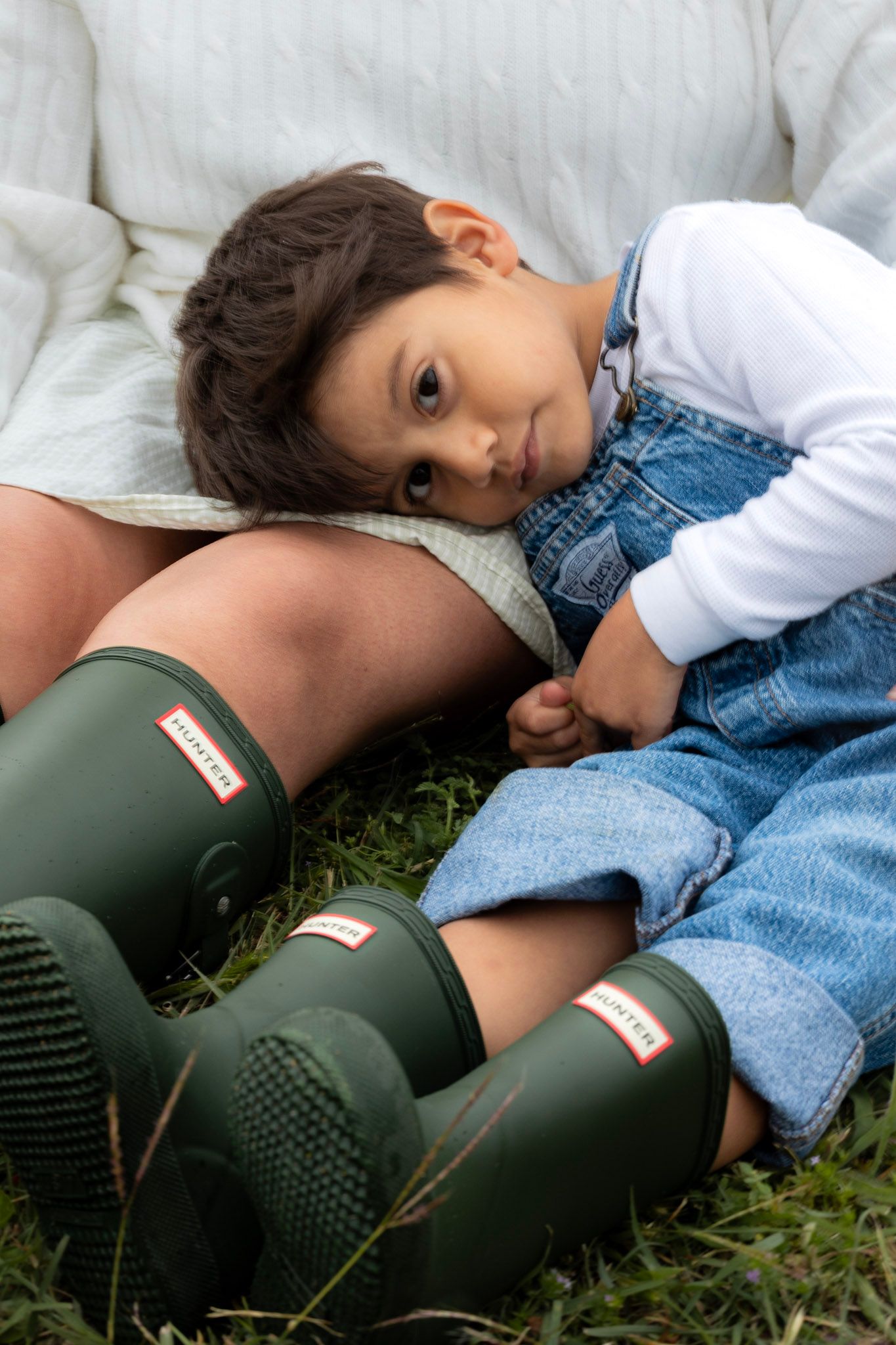 A young boy in denim overalls and white shirt rests on an adult wearing green Hunter rain boots on grass.