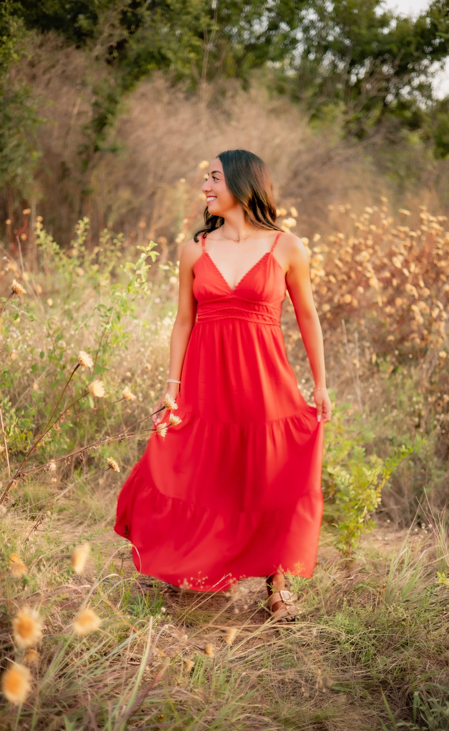 Young woman in red maxi dress walking through a sunlit meadow with wildflowers around her.