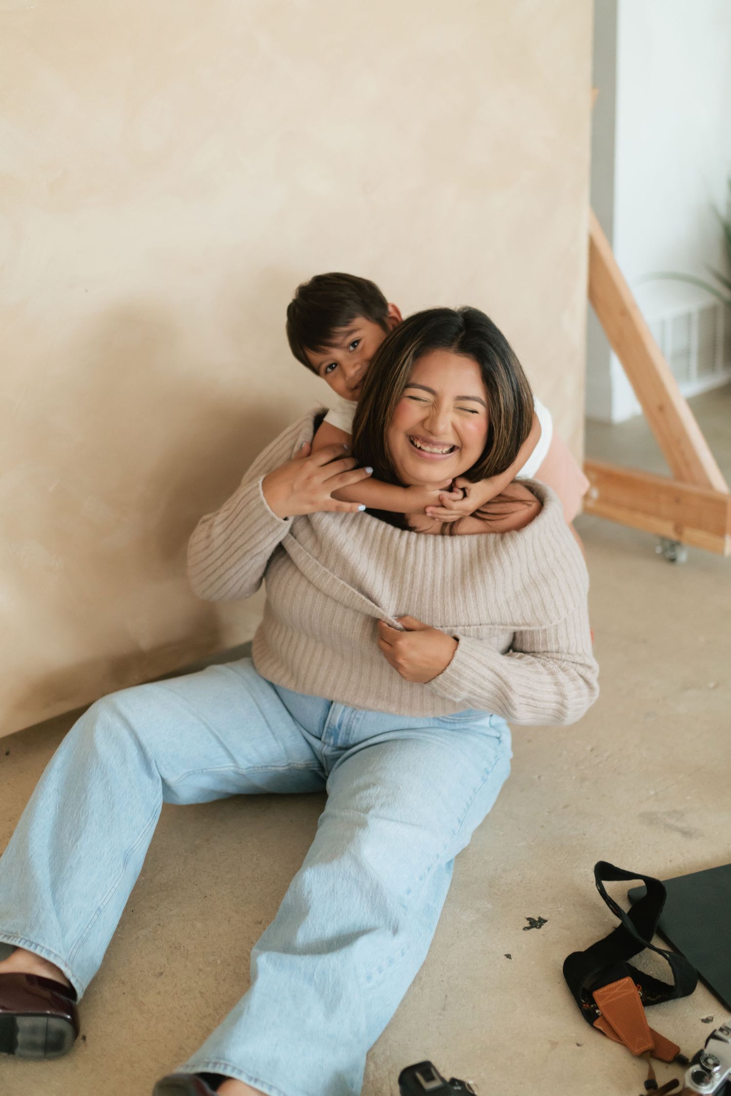Two women laughing and embracing on the floor, one hugging the other from behind in a bright, cozy indoor setting.