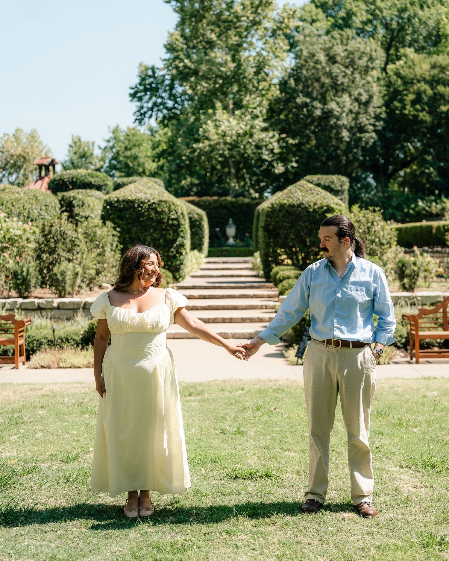 Couple holding hands in a lush garden with manicured hedges, woman in yellow dress, man in light blue shirt.