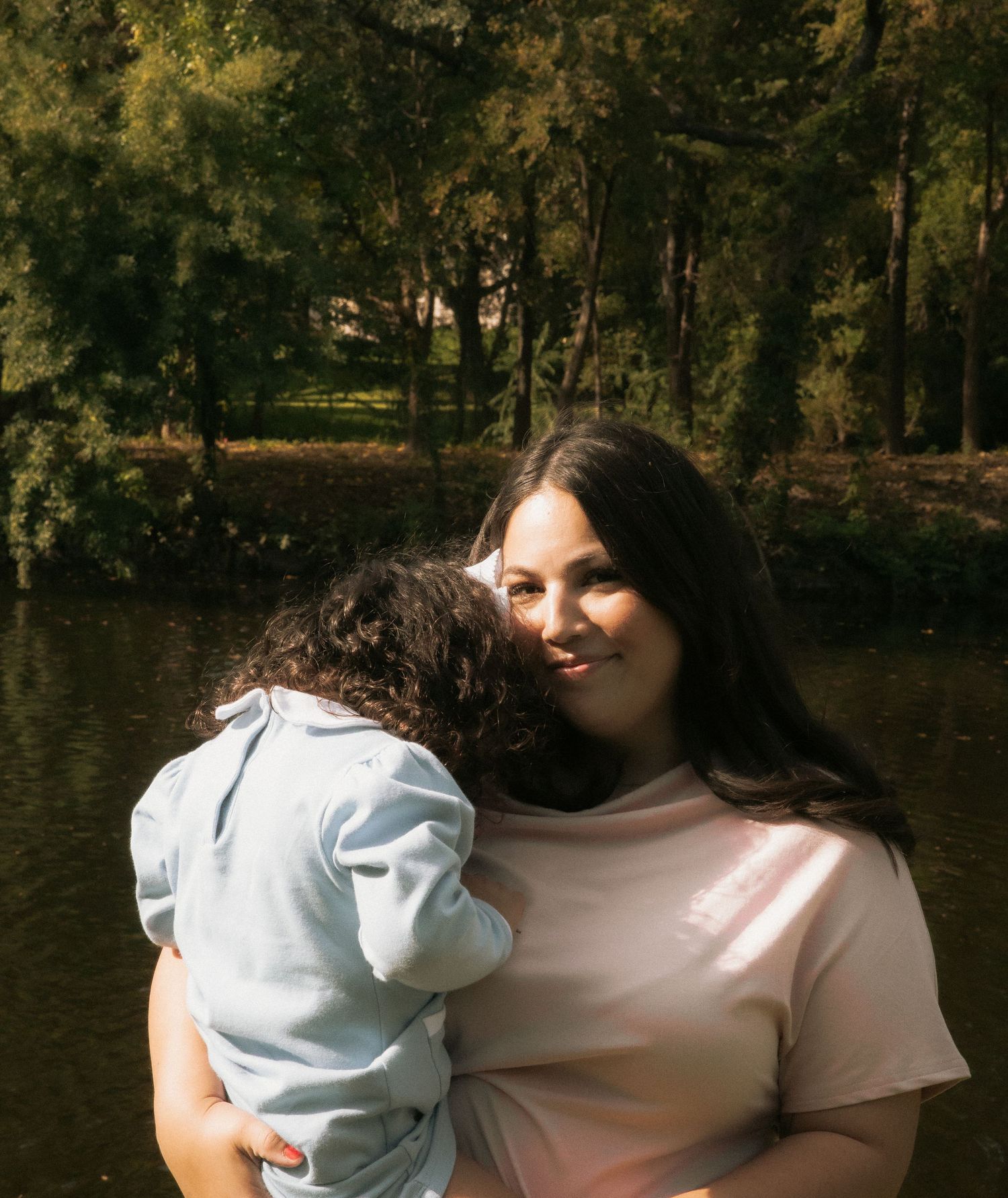 A smiling woman in a pink shirt holds a young child near a serene park pond on a sunny day.