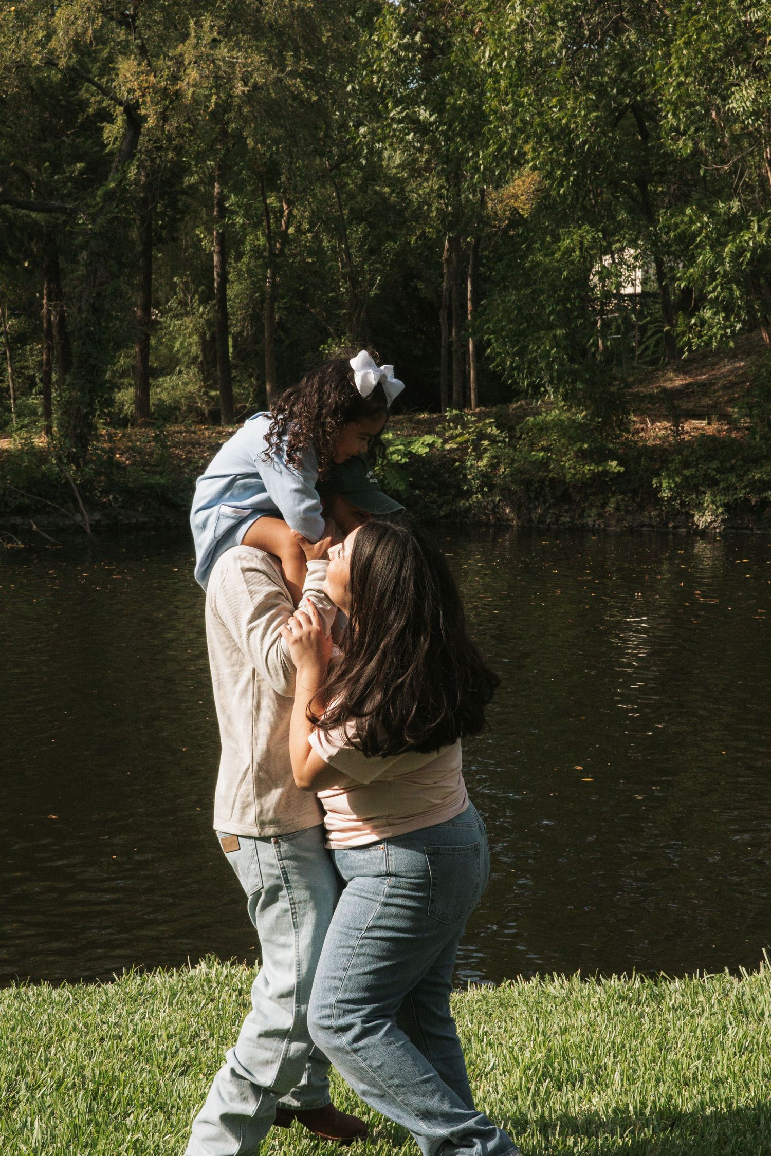 Parents lovingly lifting their young daughter outdoors near a peaceful pond surrounded by lush green trees.