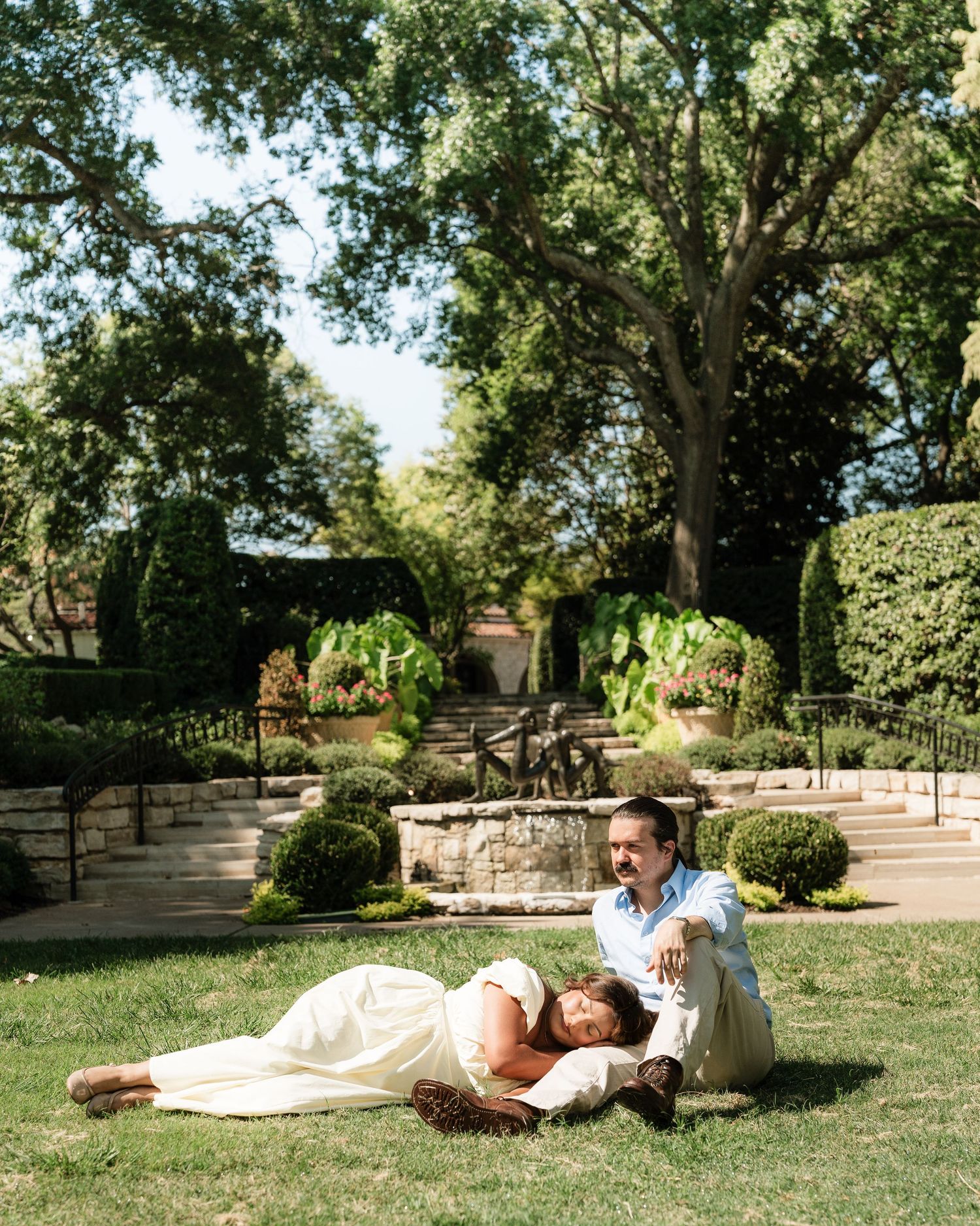 Couple relaxing on lush green grass in a beautiful garden with stone steps, hedges, and large trees in the background.