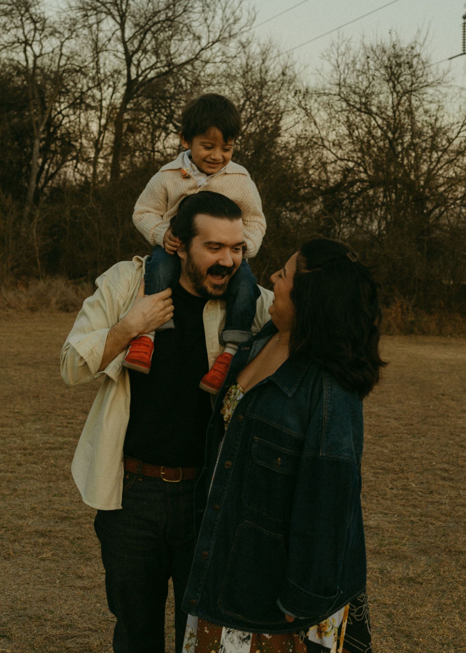 A happy family with a young child sitting on dad's shoulders while mom looks up smiling in an outdoor setting.