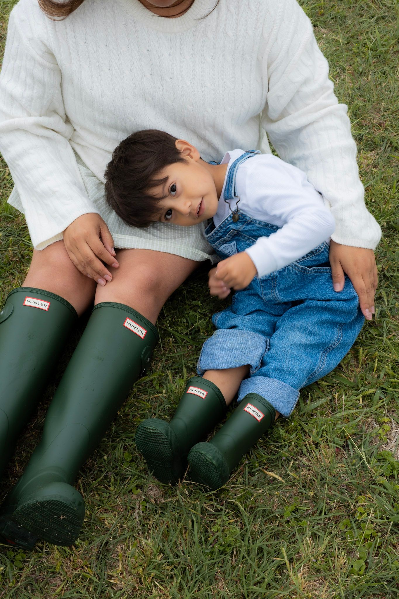 Child in denim overalls leaning on adult wearing white sweater and green Hunter rain boots, sitting on grass.