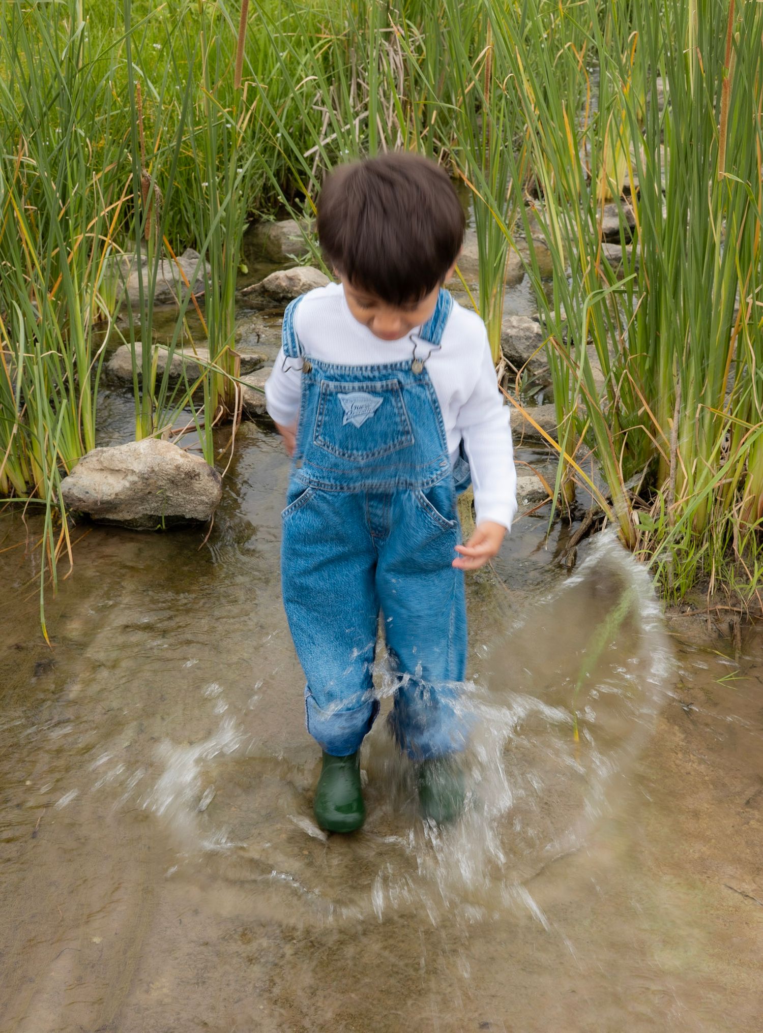 Young child in denim overalls and green boots splashing through a shallow stream outdoors.