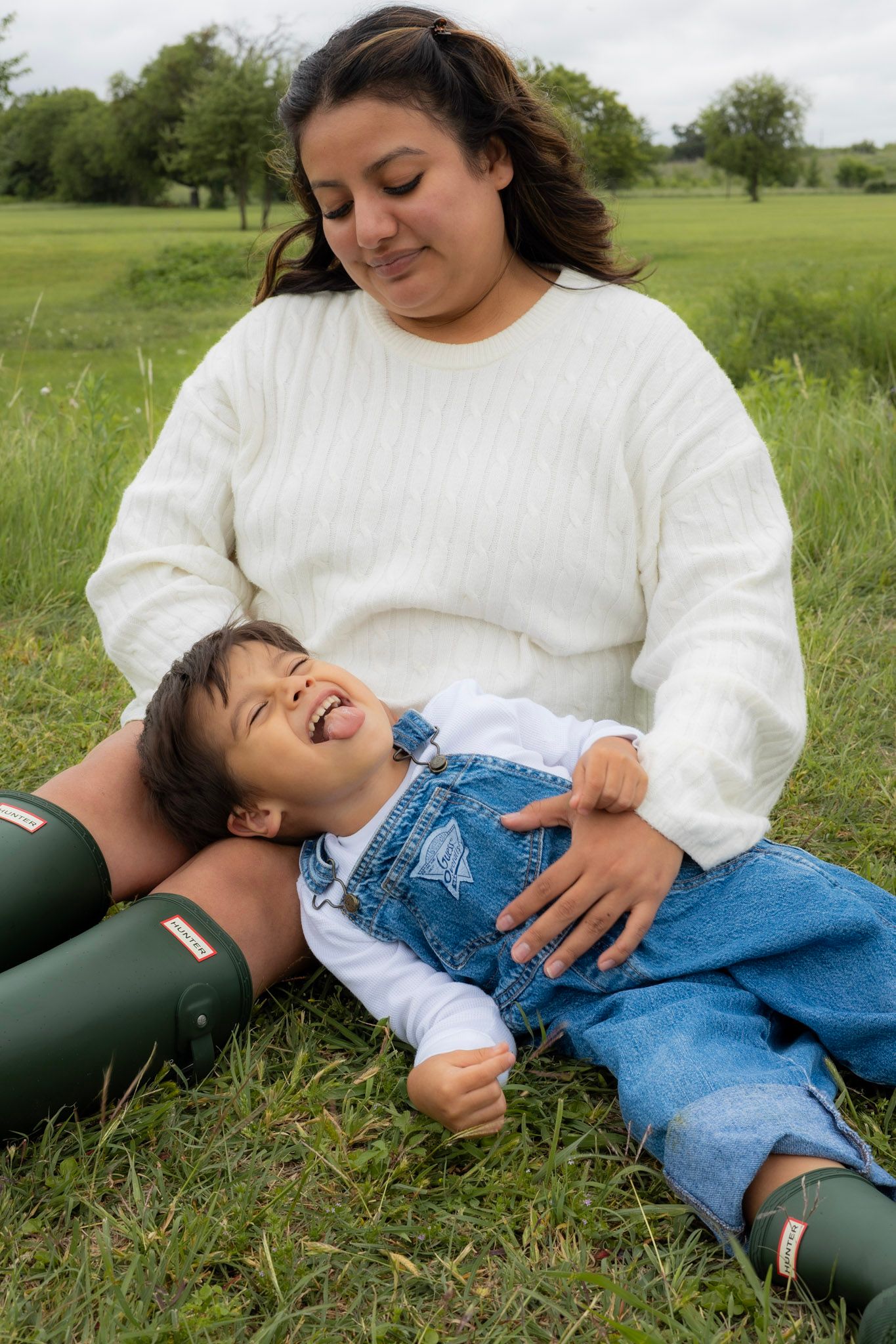 A mother in a white sweater sits on grass laughing with her young child in denim overalls and green Hunter boots.