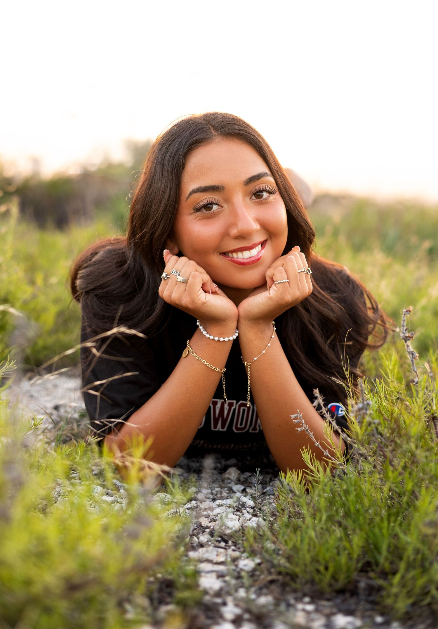 Smiling young woman lying in green grass wearing a black TWU shirt with jewelry at sunset.