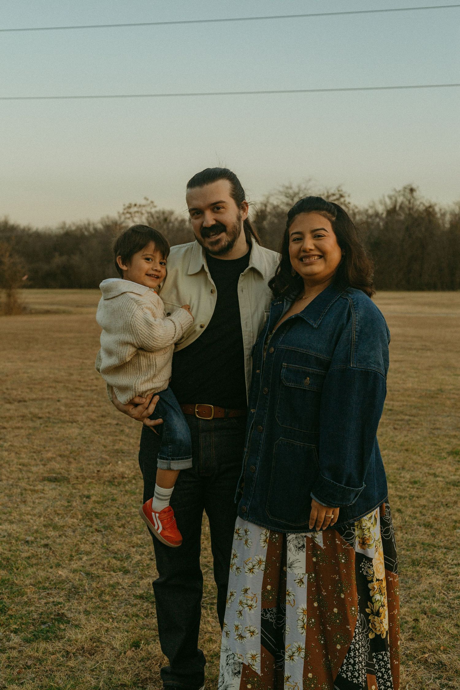 A smiling family of three poses outdoors at dusk, father holding toddler beside mother in floral skirt on grassy field.