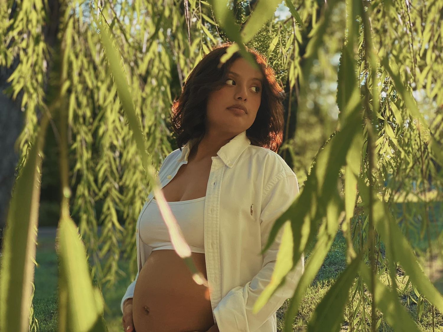 Young woman in white crop top and open shirt standing among weeping willow branches in golden sunlight.