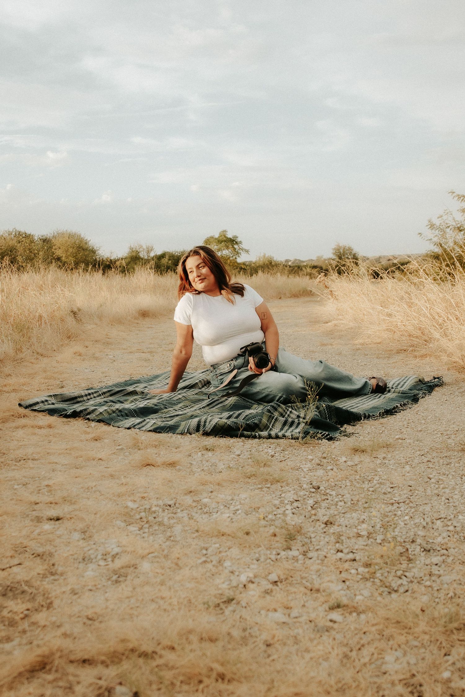 Woman in white top and jeans sitting on a patterned blanket in a dry grassy field under a cloudy sky.