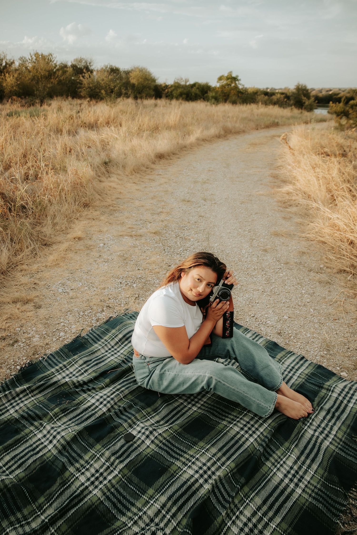 Smiling woman in white tee hugs camera while sitting barefoot on plaid blanket on rural dirt path.