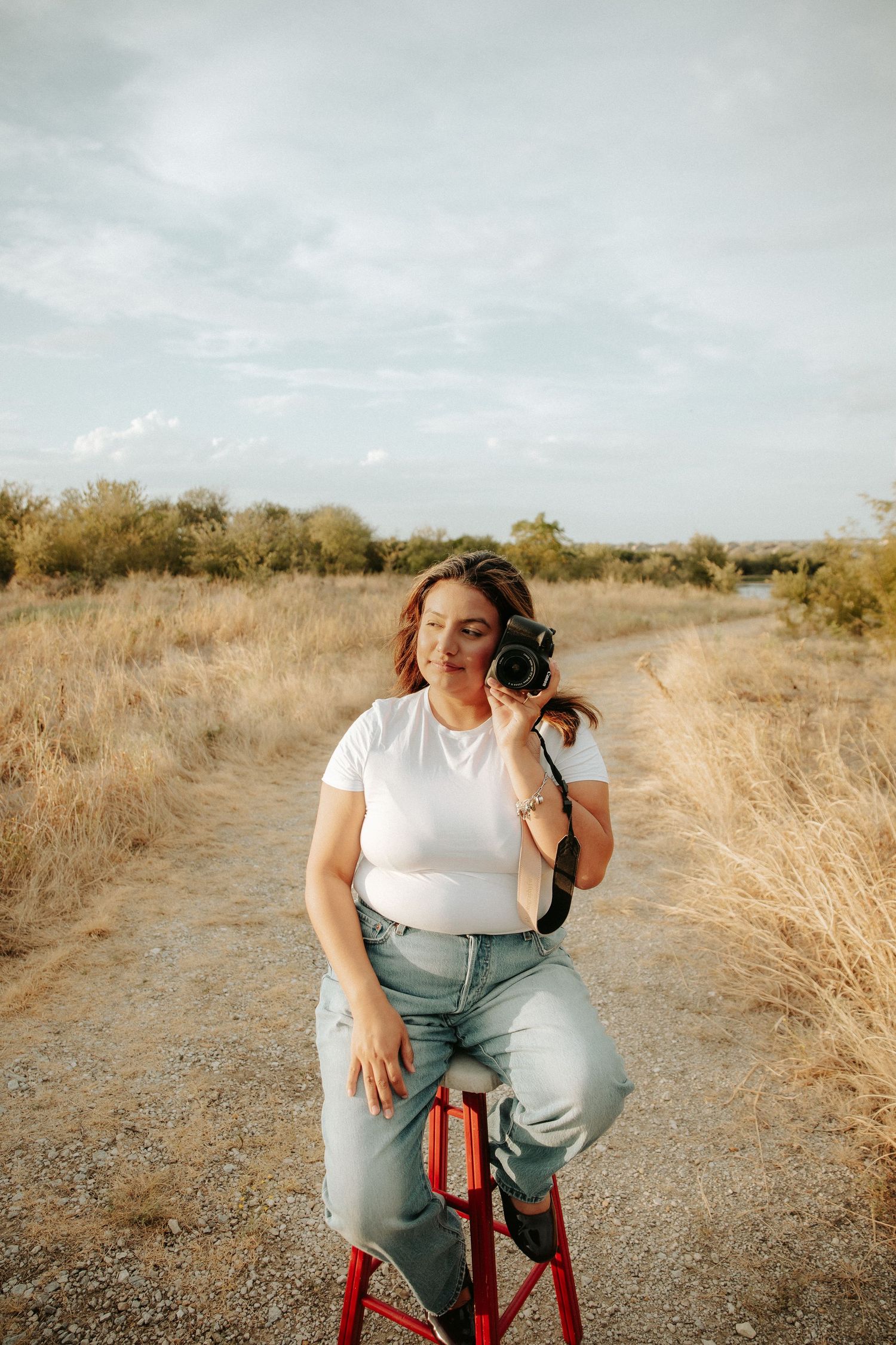 Woman in white tee and jeans sits on red stool on dirt path holding camera in golden field.