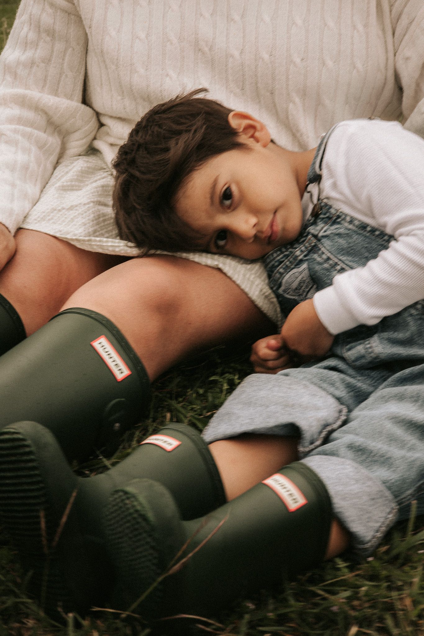 Child in denim overalls resting on adult wearing Hunter green rain boots, sitting together on grass outdoors.