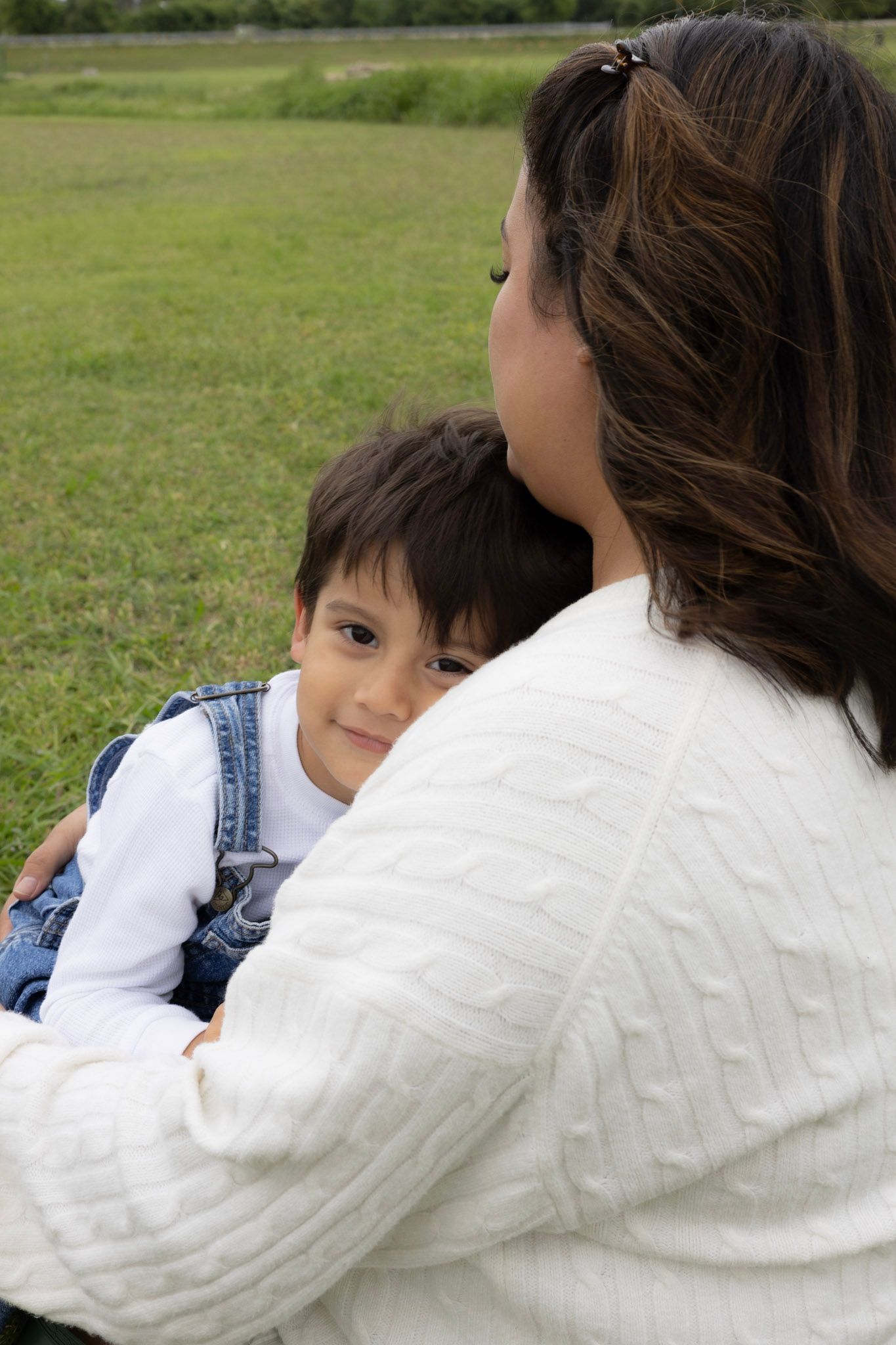 A mother holds her young boy in her arms outdoors in a green field, as he looks up at her with a gentle expression.