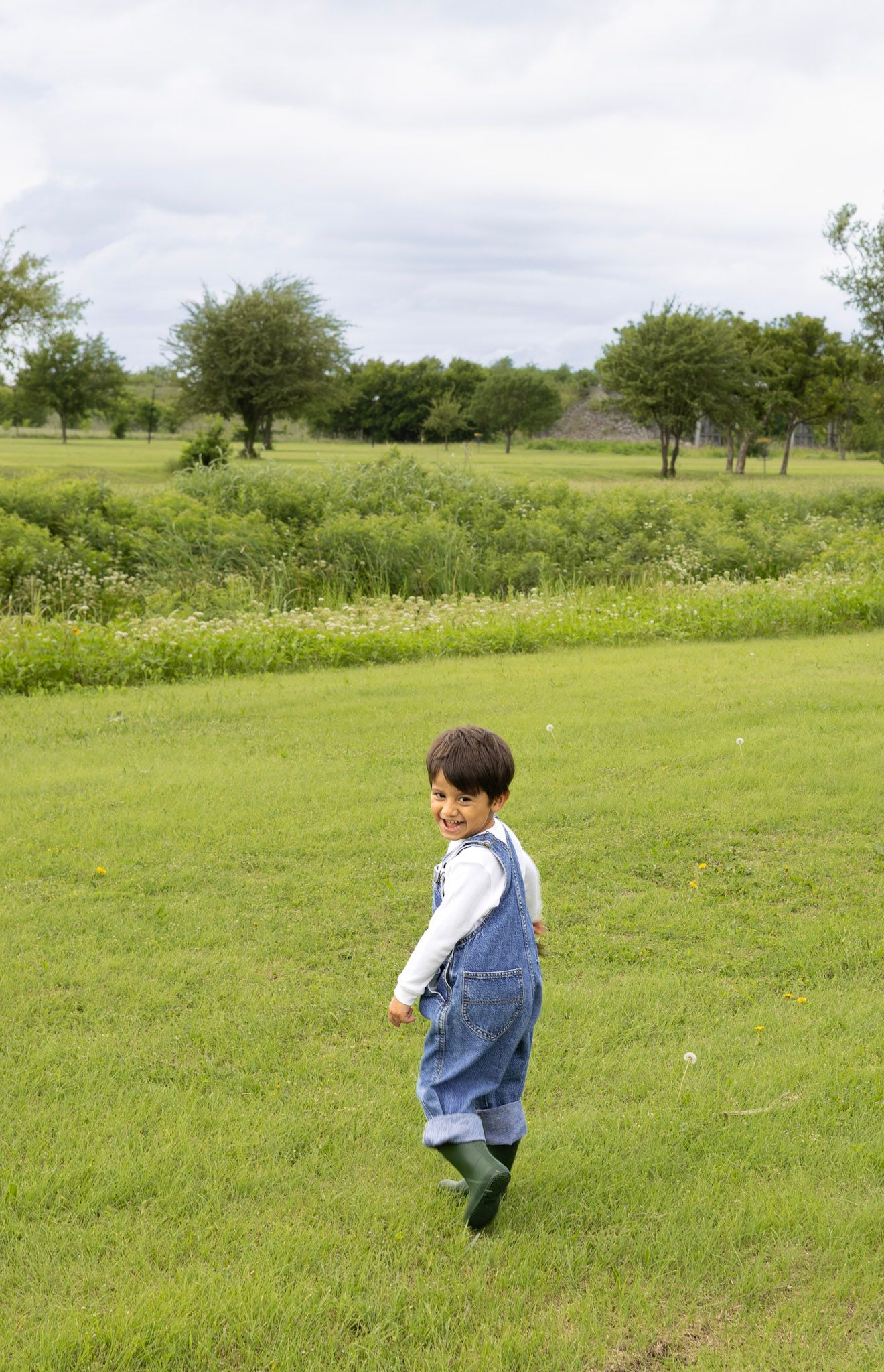 Smiling toddler in denim overalls and green rain boots playing in a green field with trees.