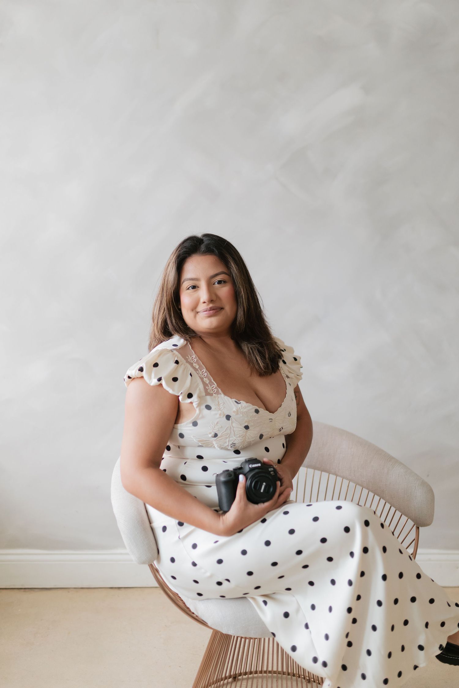 Female photographer in white polka dot dress sitting in chair holding camera indoors.