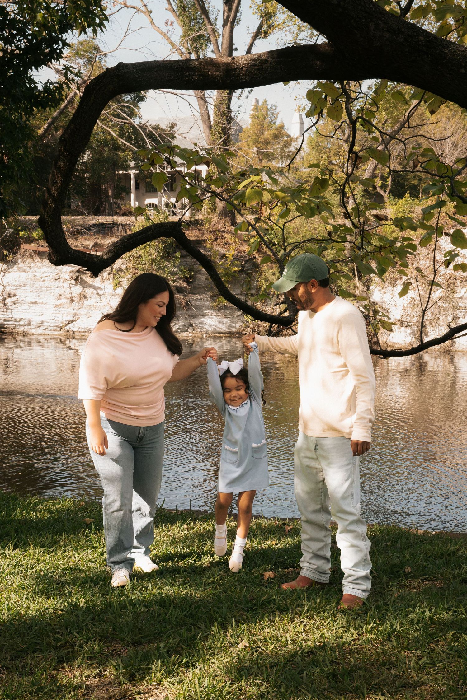 Family of three playing outdoors near a pond, parents holding toddler's hands under a tree on a sunny day.