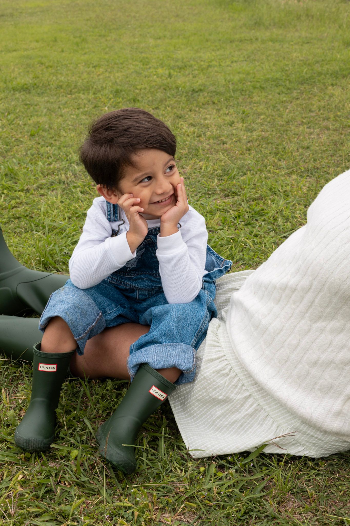 Smiling toddler in denim overalls and green Hunter boots sitting on grass with hands on cheeks.