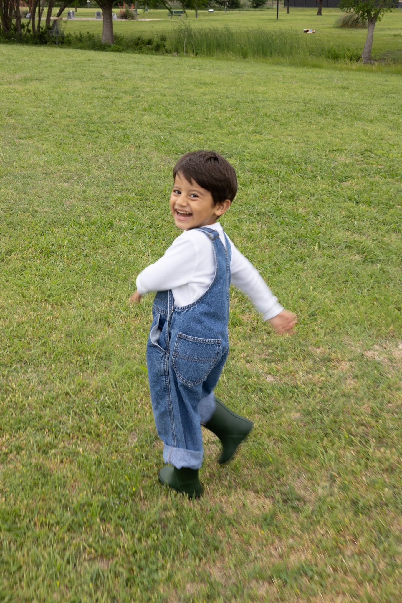 Cheerful toddler boy in denim overalls and green boots running and smiling in a green park.