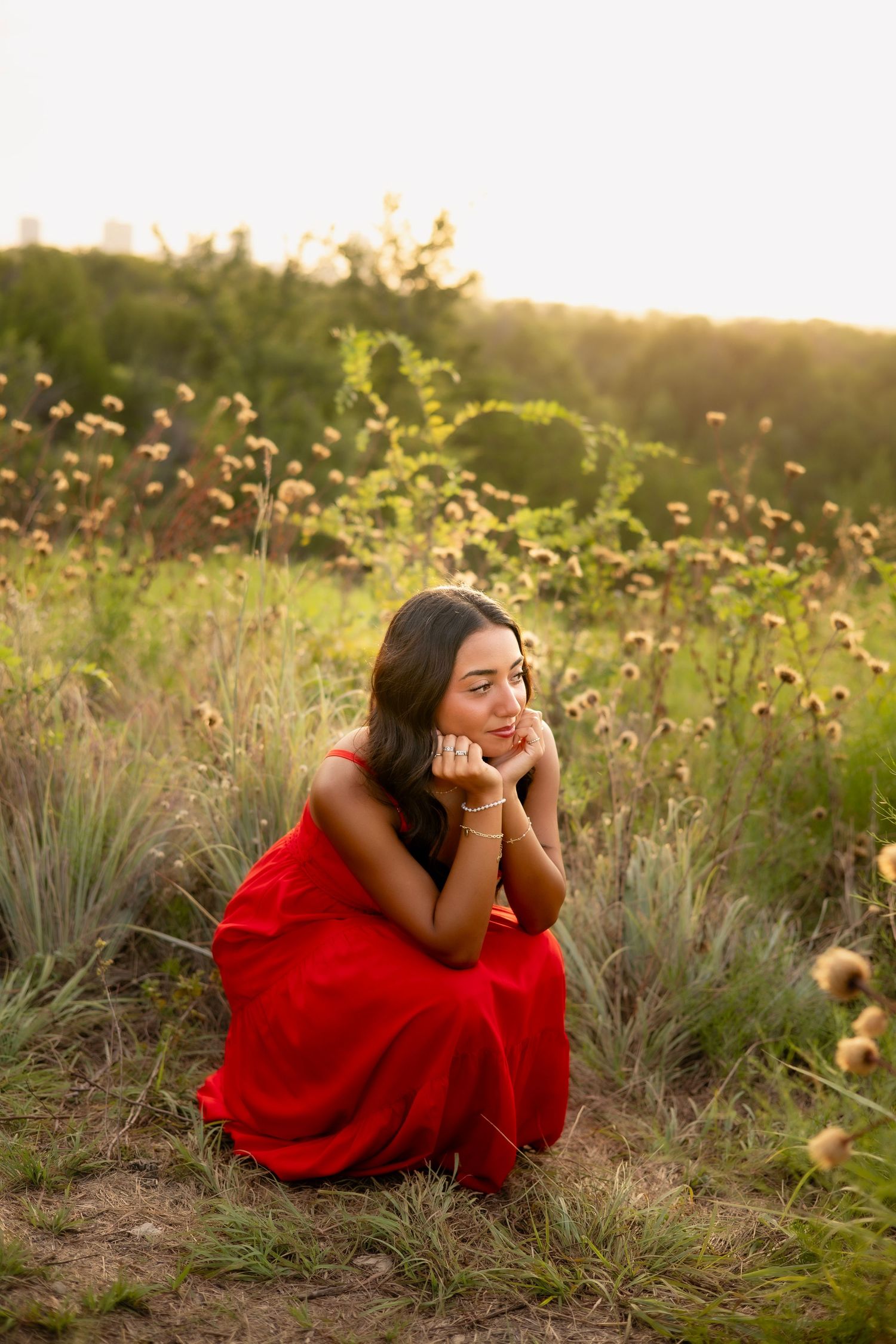 Young woman in red dress crouching thoughtfully among wildflowers in a golden hour meadow.
