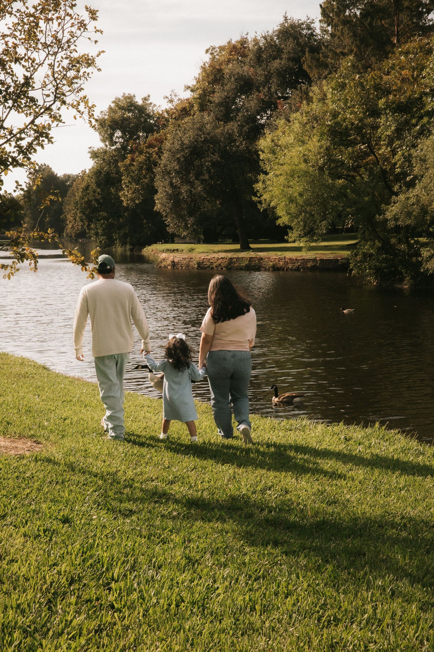 Family of three walking hand-in-hand along a serene park lake surrounded by lush green trees.
