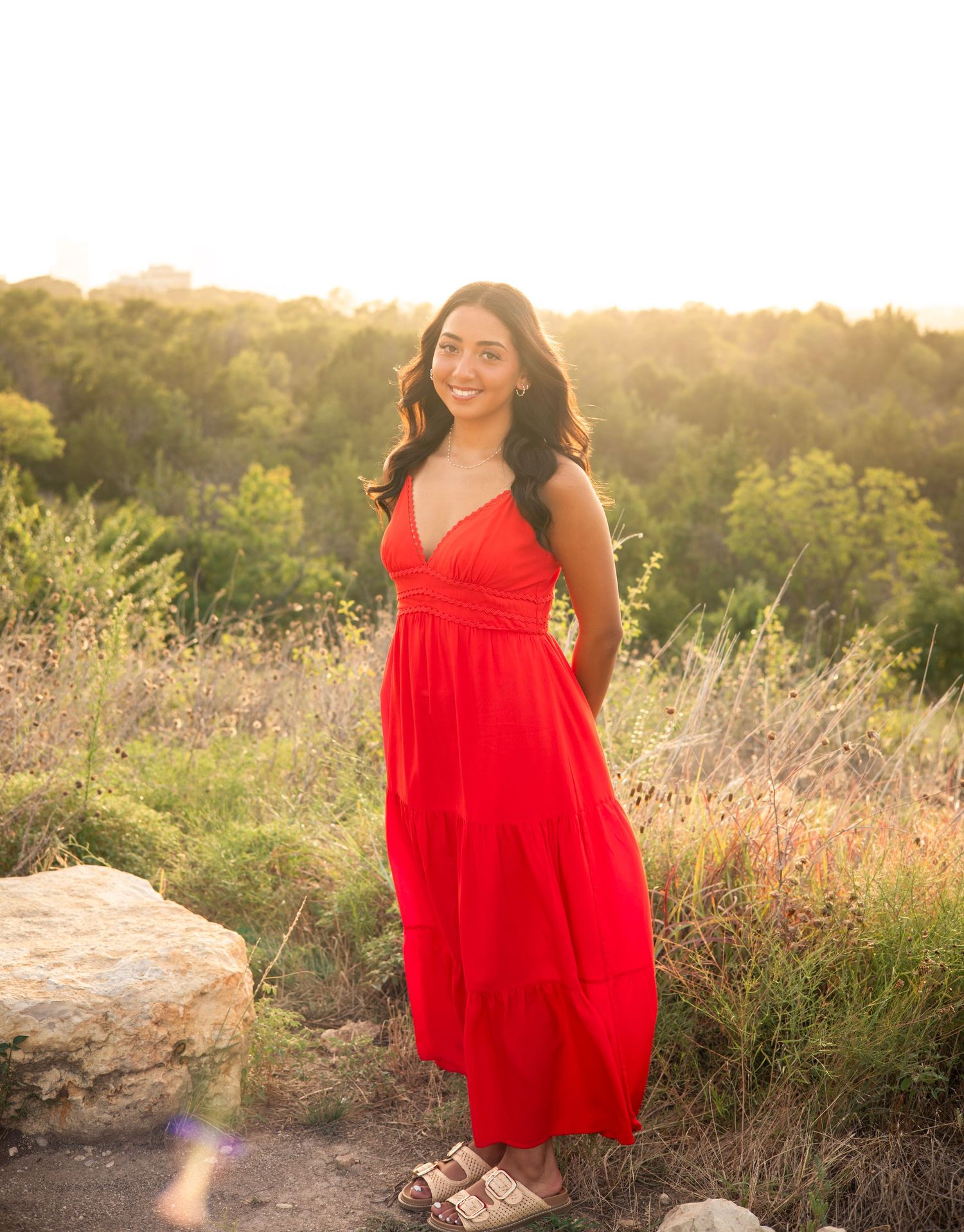 Young woman in red maxi dress smiling outdoors at golden hour in a natural field setting.