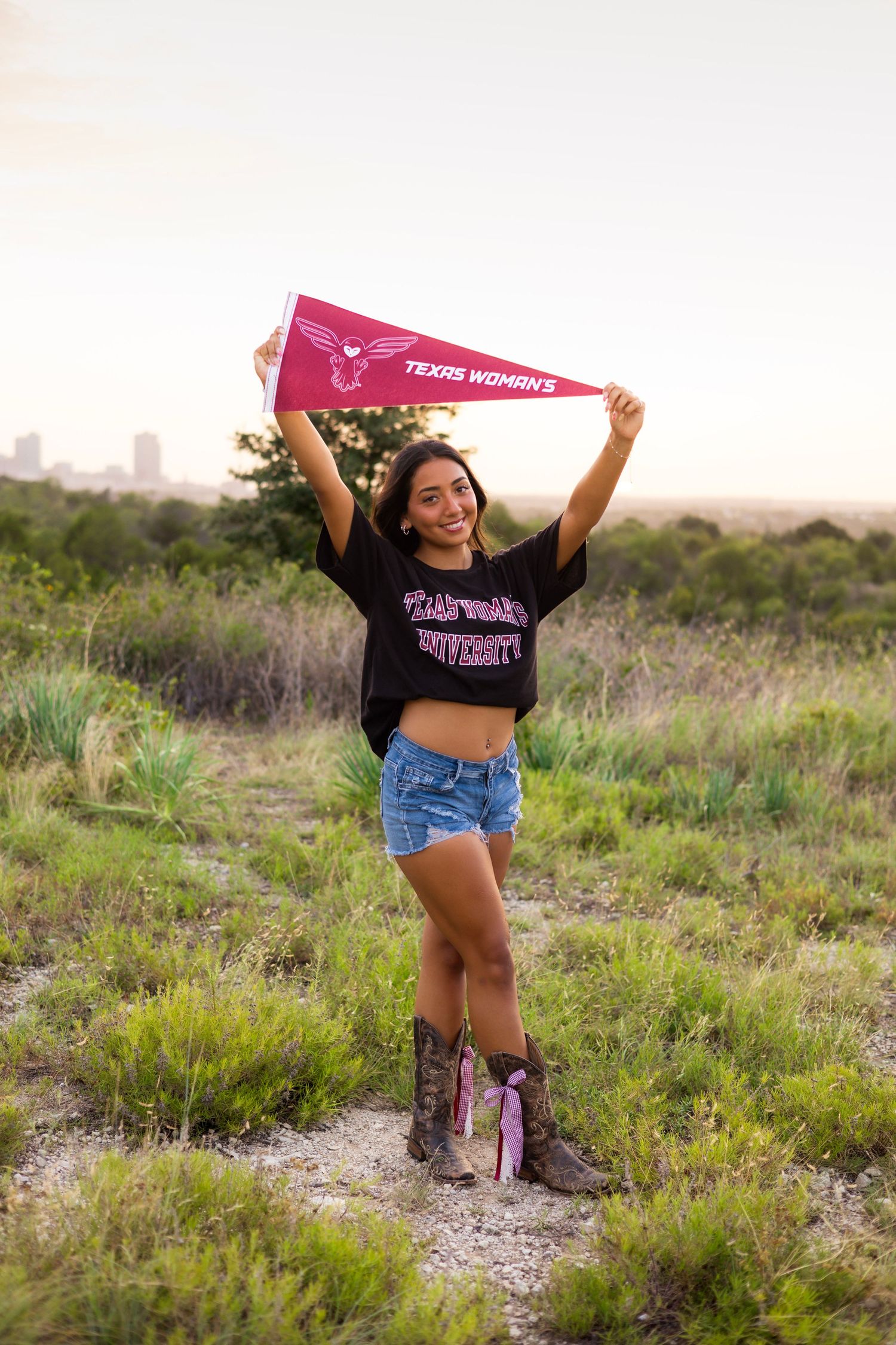 Smiling young woman in TWU shirt and denim shorts holding Texas Woman's University pennant overhead.