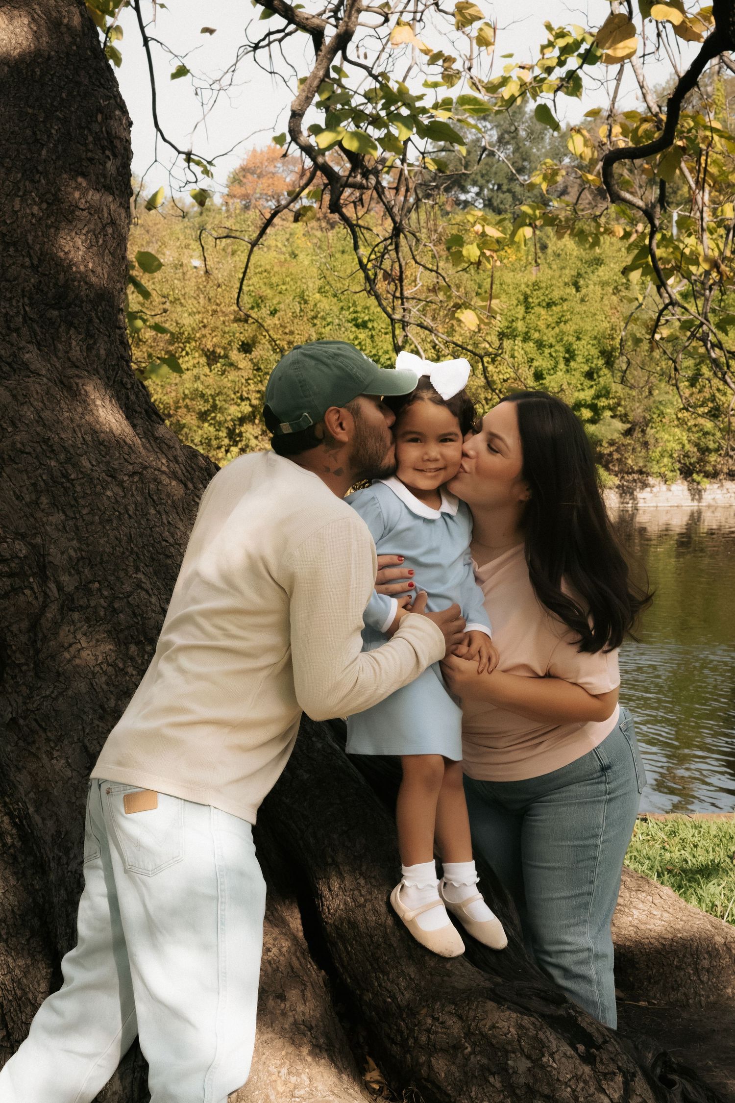 Parents kissing their toddler daughter on the cheeks while sitting on a tree root in a park.